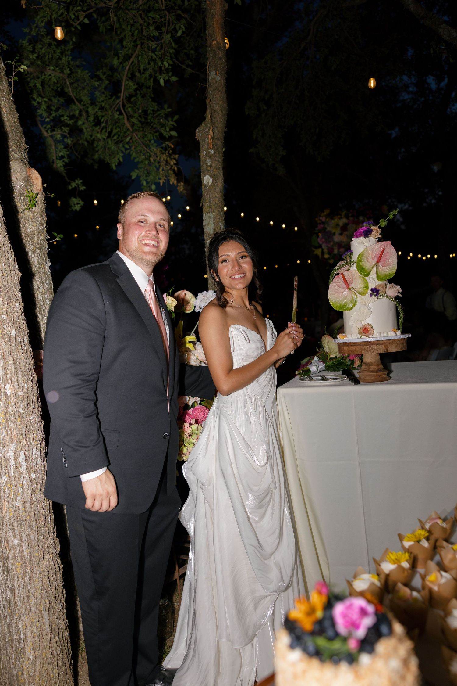 Bride and groom smiling together at outdoor wedding reception beside elegant floral cake at night.