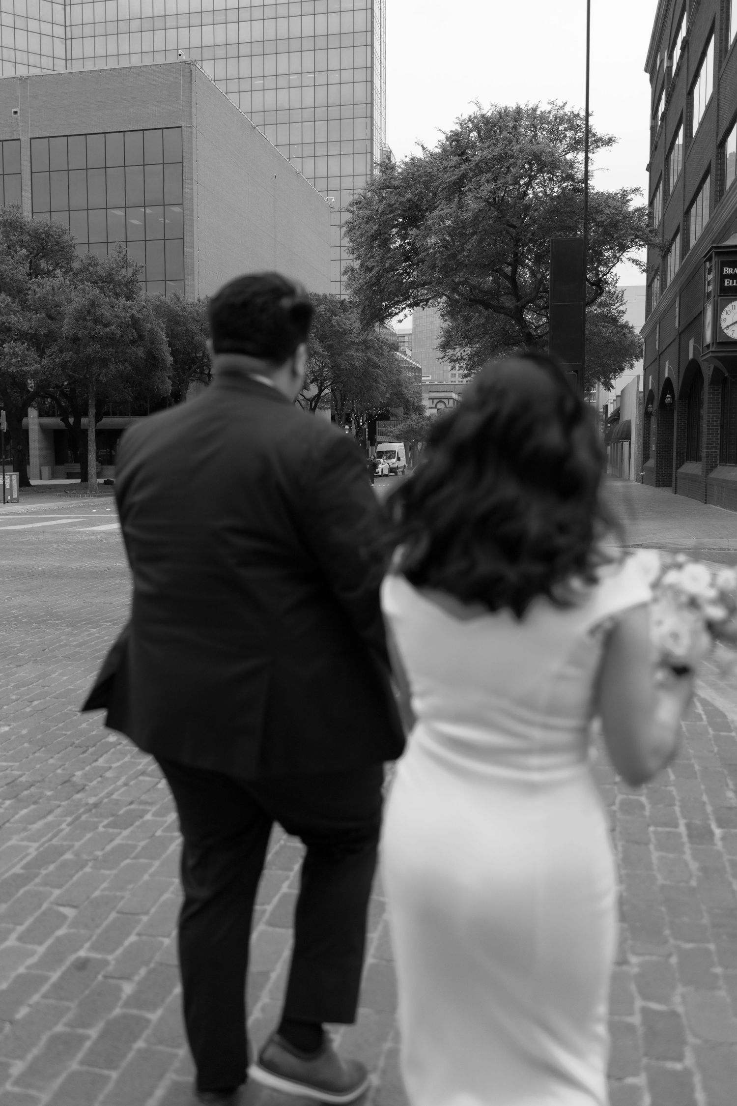 Black and white photo of a bride and groom walking away on a cobblestone street in an urban setting.