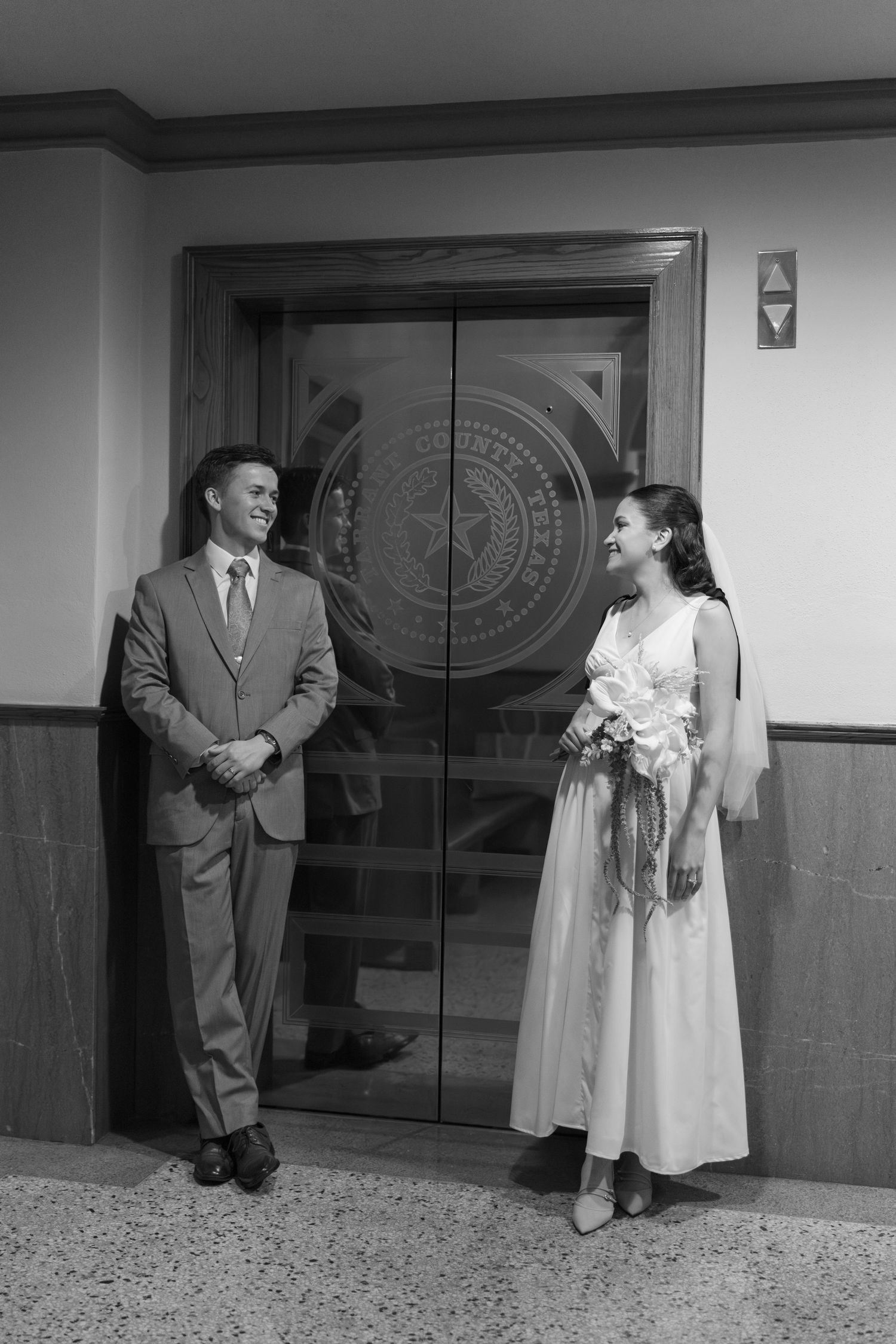 Bride and groom smiling at each other in black and white photo beside elevator with Texas state seal.