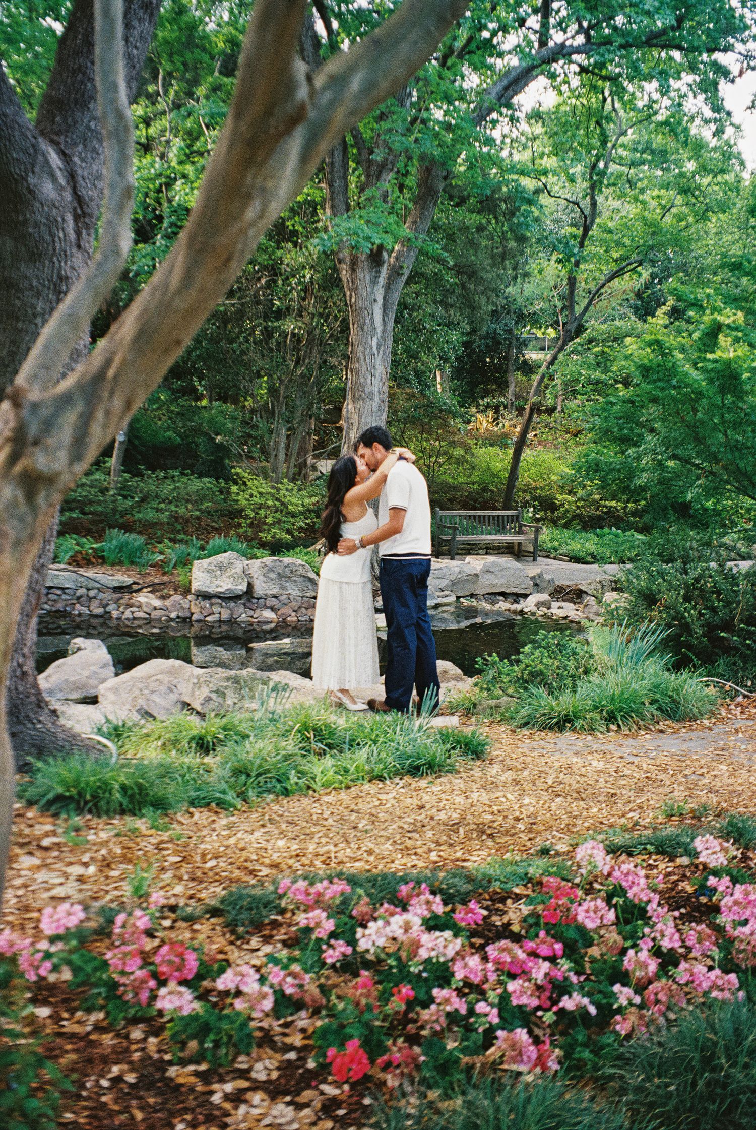 Couple sharing a romantic kiss in a lush garden surrounded by pink flowers, rocks, and green trees.