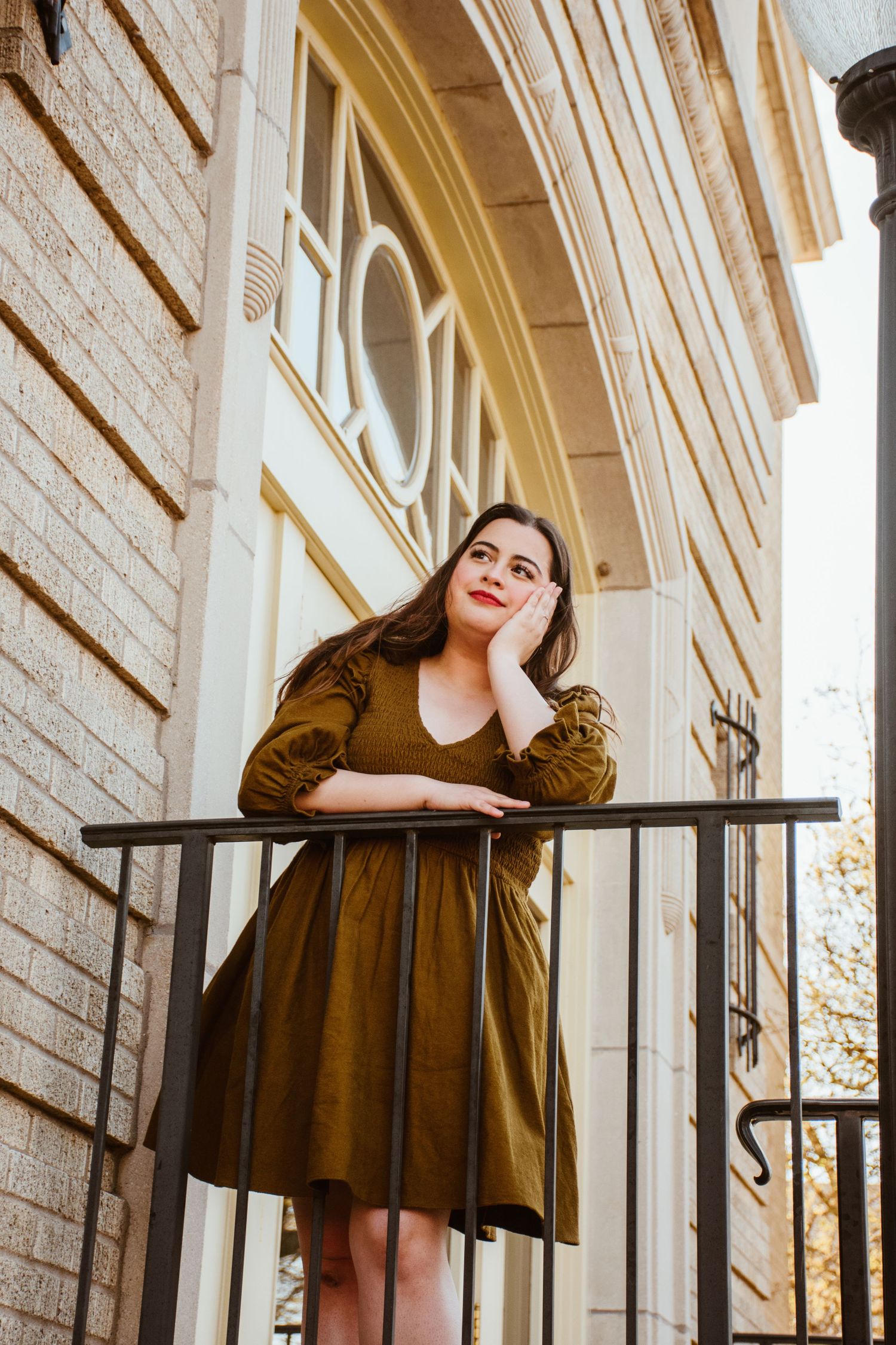 Woman in brown dress poses against white brick wall and arched windows in outdoor fashion photoshoot.
