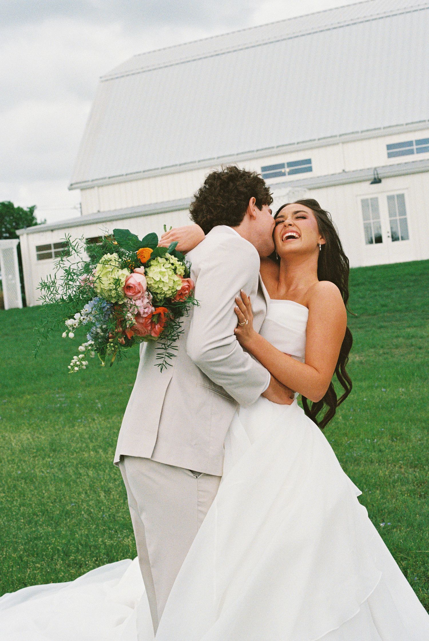 Bride laughing joyfully as groom kisses her cheek outdoors, holding colorful wedding bouquet with white barn backdrop.