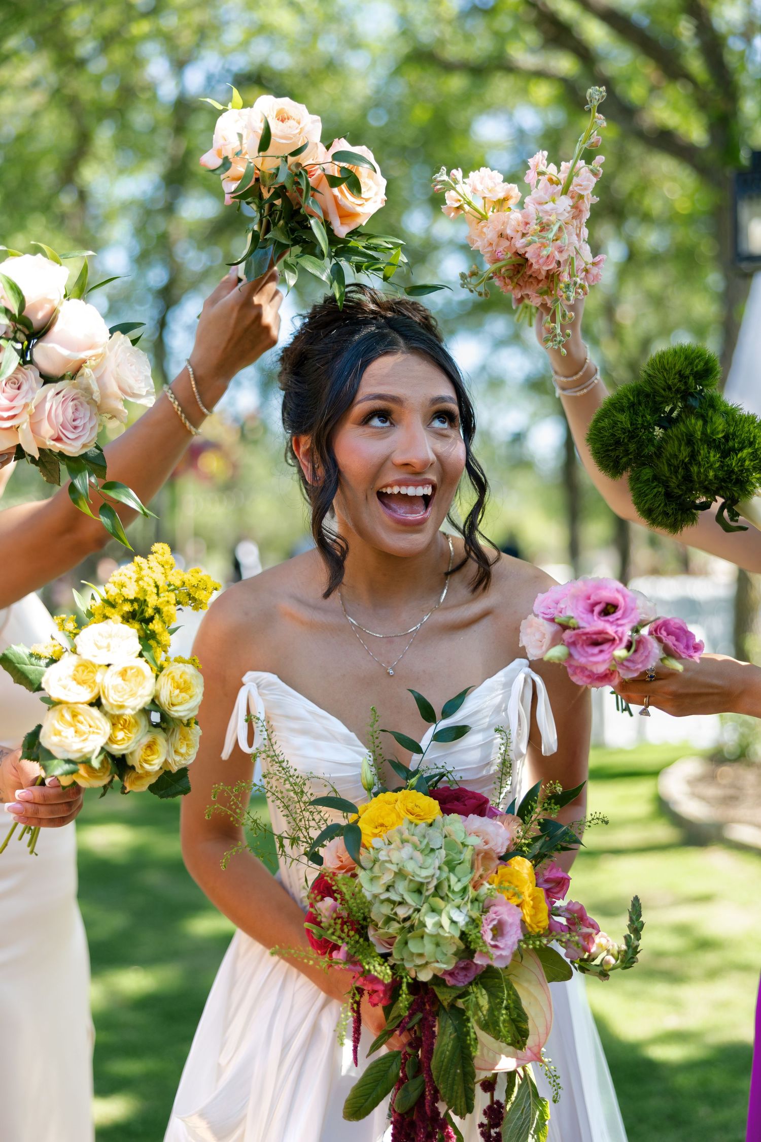 Joyful bride in white gown laughs as bridesmaids raise colorful bouquets in sunny outdoor setting.