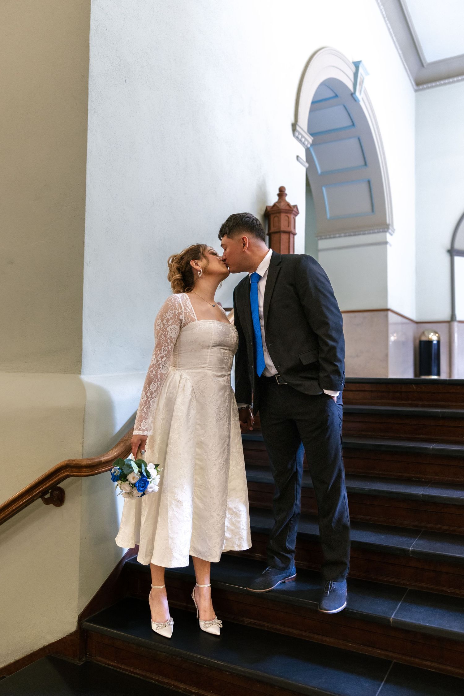 Bride and groom share a kiss on elegant staircase, bride holding blue bouquet in white midi dress.