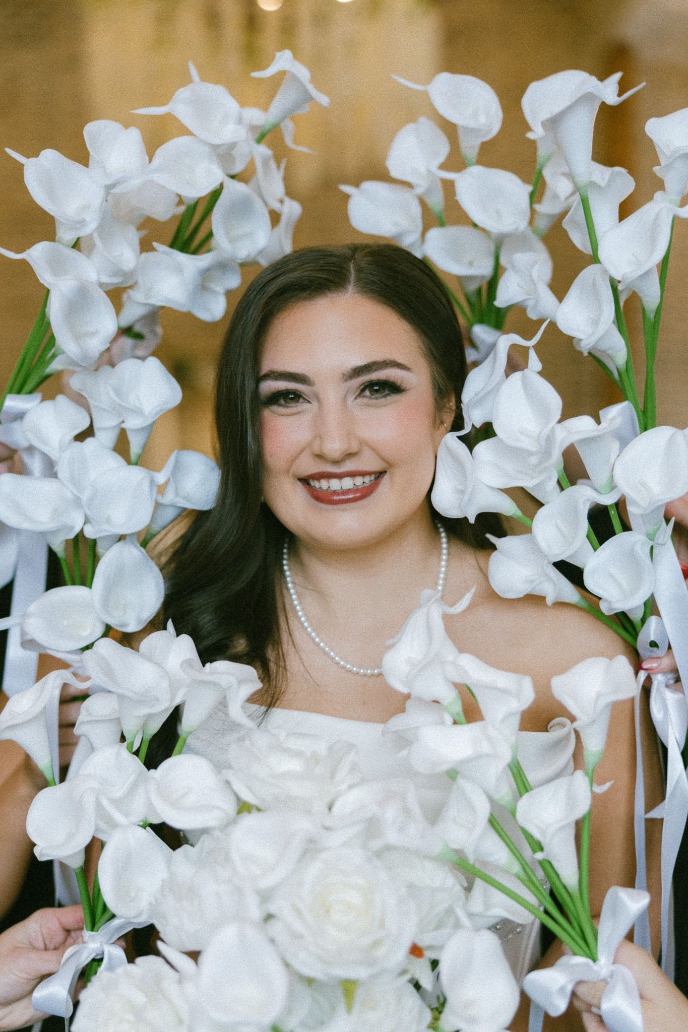 Smiling bride in white dress and pearl necklace surrounded by elegant white calla lily flowers holding a bouquet.