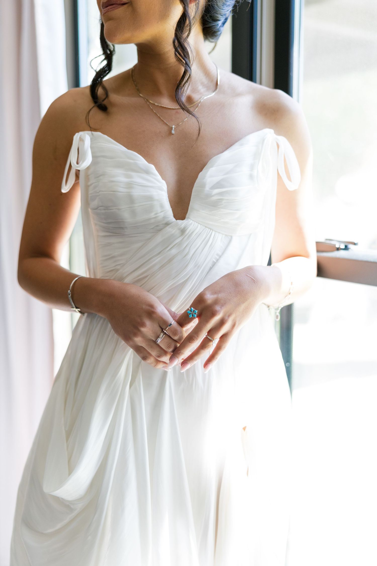 Bride in flowing white strapless wedding gown with deep V-neckline, wearing delicate jewelry near window.