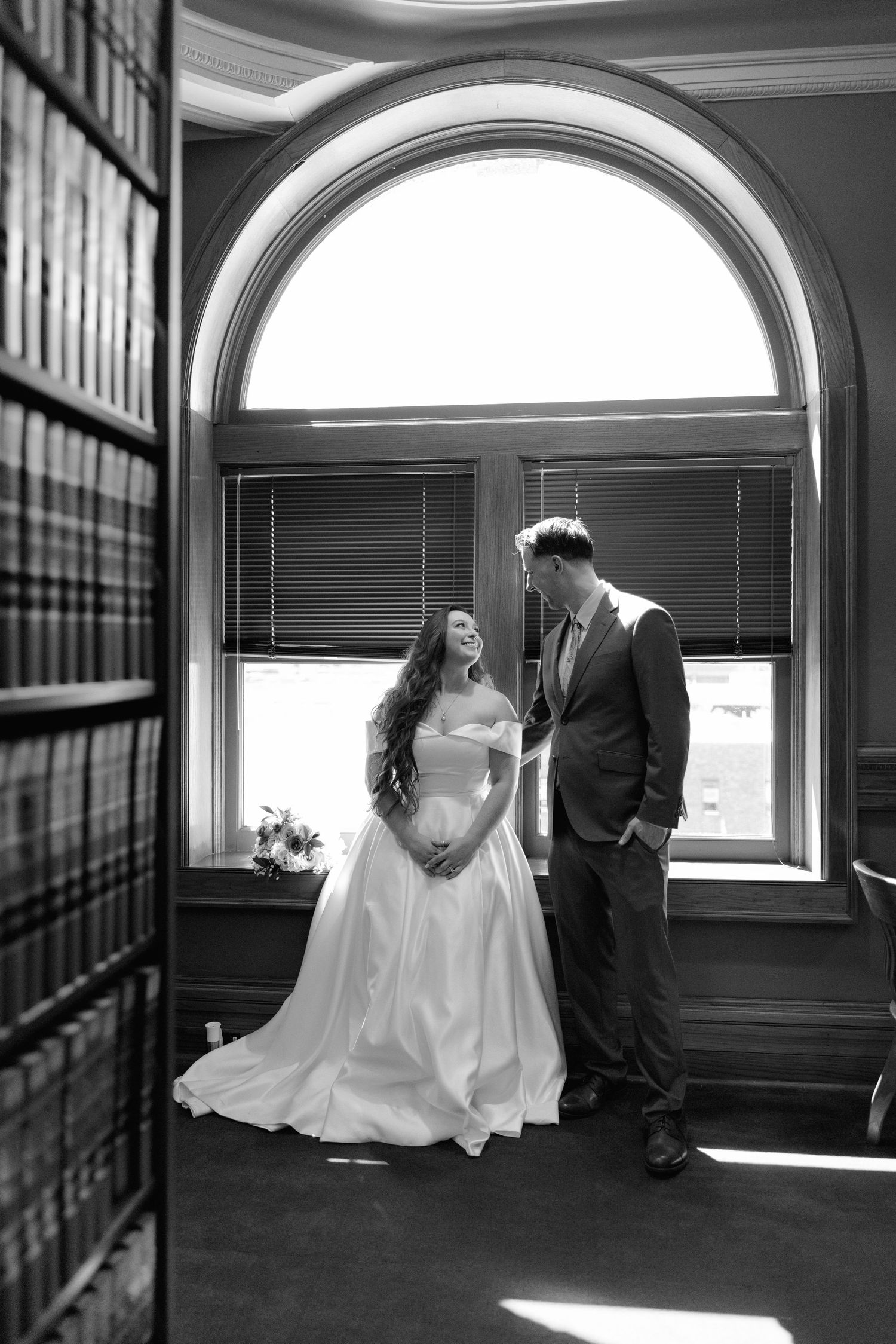 Black and white photo of bride and groom sharing a tender moment in a sunlit library with arched window.