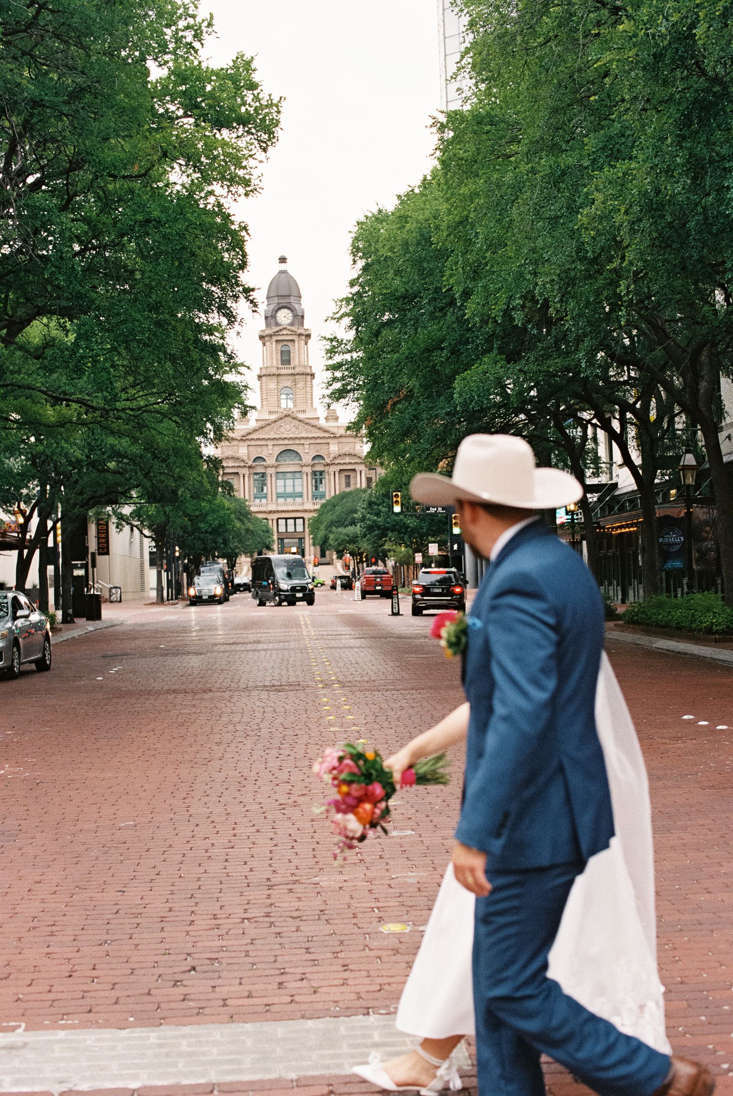 Bride and groom in blue suit and cowboy hat walk down brick street with bouquet, historic courthouse in background.