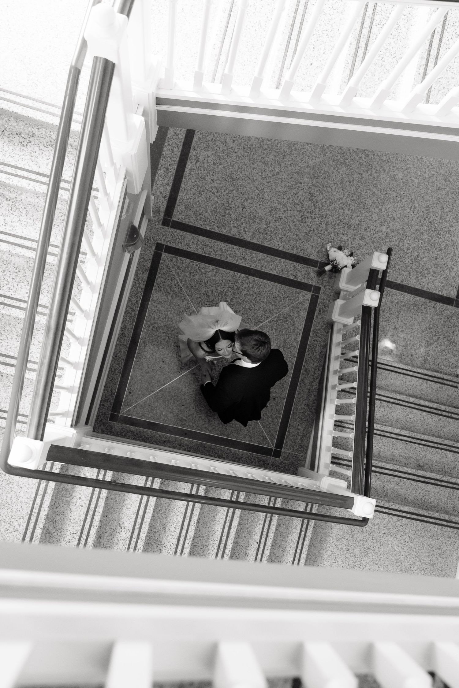 Aerial black and white photo of a bride and groom embracing at the bottom of an elegant stairwell.