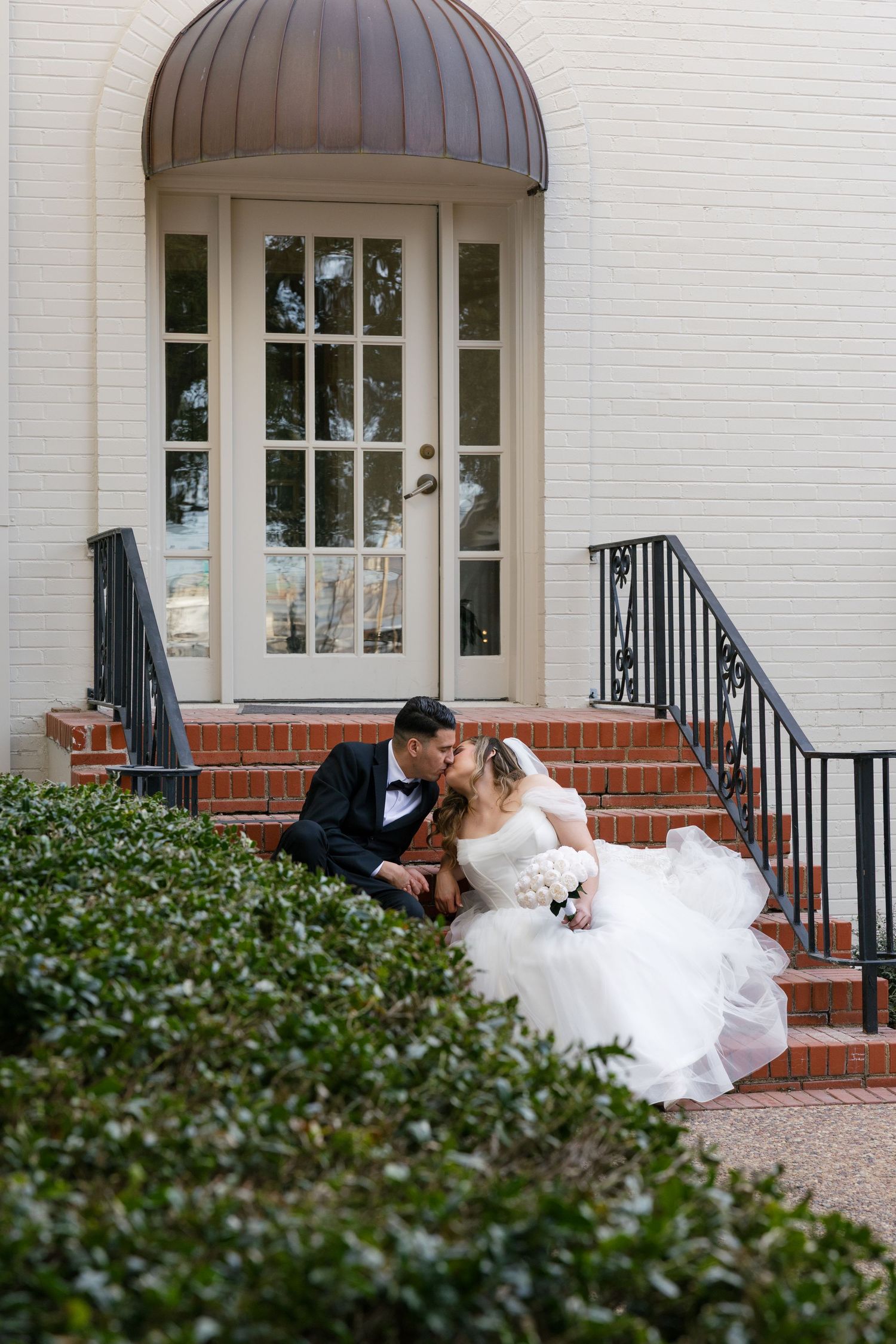 Bride and groom share a romantic kiss on brick steps outside a white building with an elegant arched doorway.