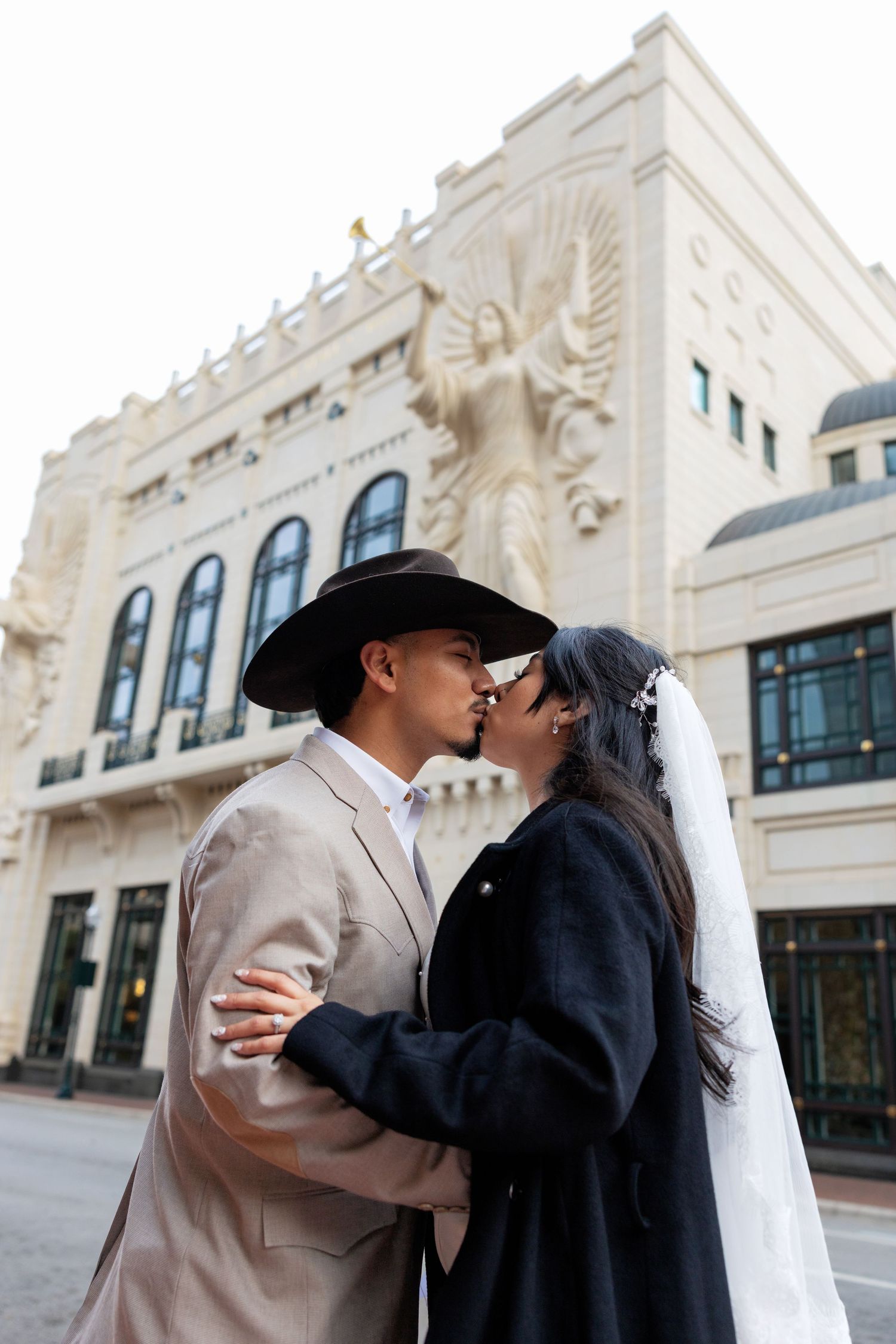A couple in formal attire share a romantic kiss outside a historic building with ornate architecture.