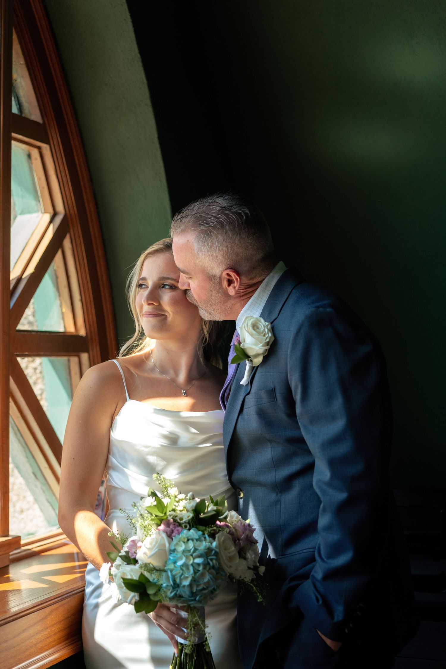 A tender wedding moment by a window with natural light streaming in, featuring a white dress and navy suit.
