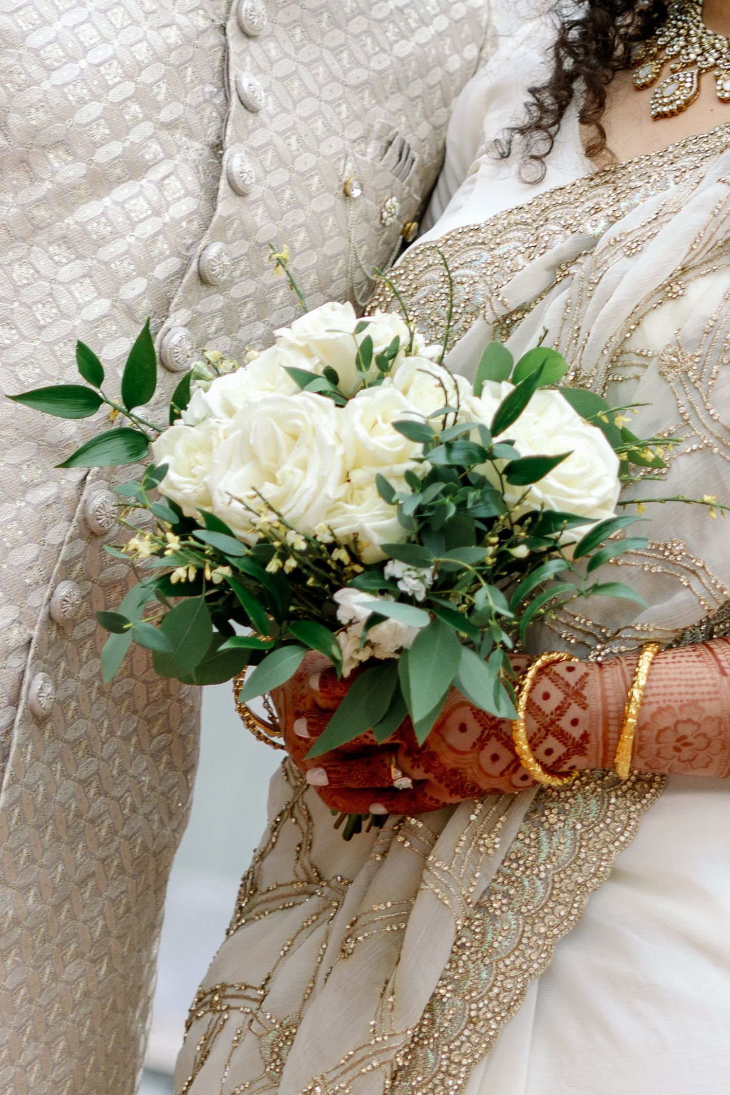 Bride in gold embroidered sari holding white rose bouquet with greenery, gold bangles visible, standing beside groom.