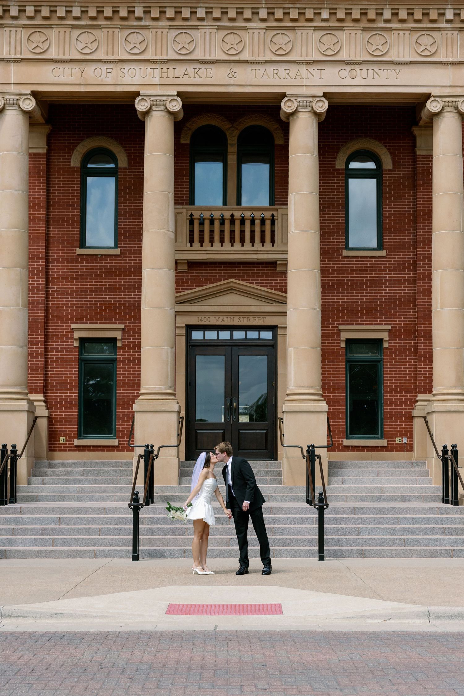 Bride and groom kissing in front of City of Southlake and Tarrant County brick building with columns.