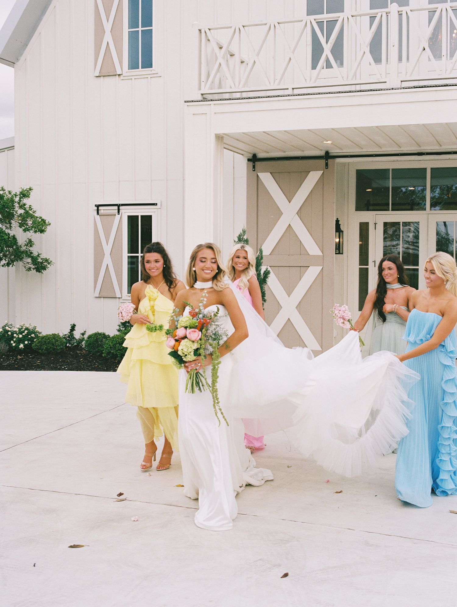 Bride in white gown walks with bridesmaids in pastel dresses outside a white barn wedding venue.