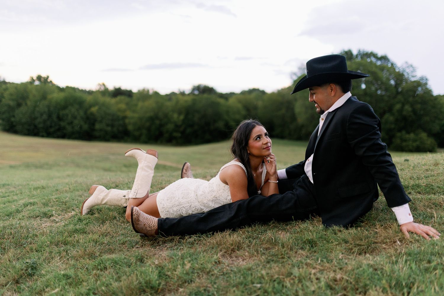 Bride and groom relaxing on green grass field, she in lace dress with boots, he in black tuxedo and cowboy hat.