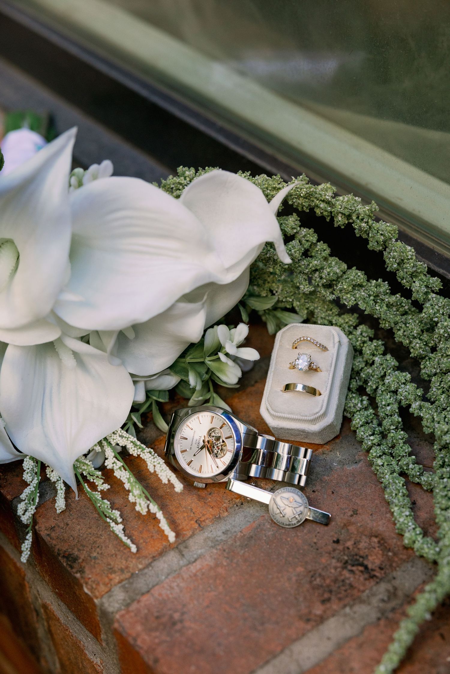Wedding rings in gold box beside a mechanical watch, cufflinks, white calla lilies, and green succulents on brick surface.