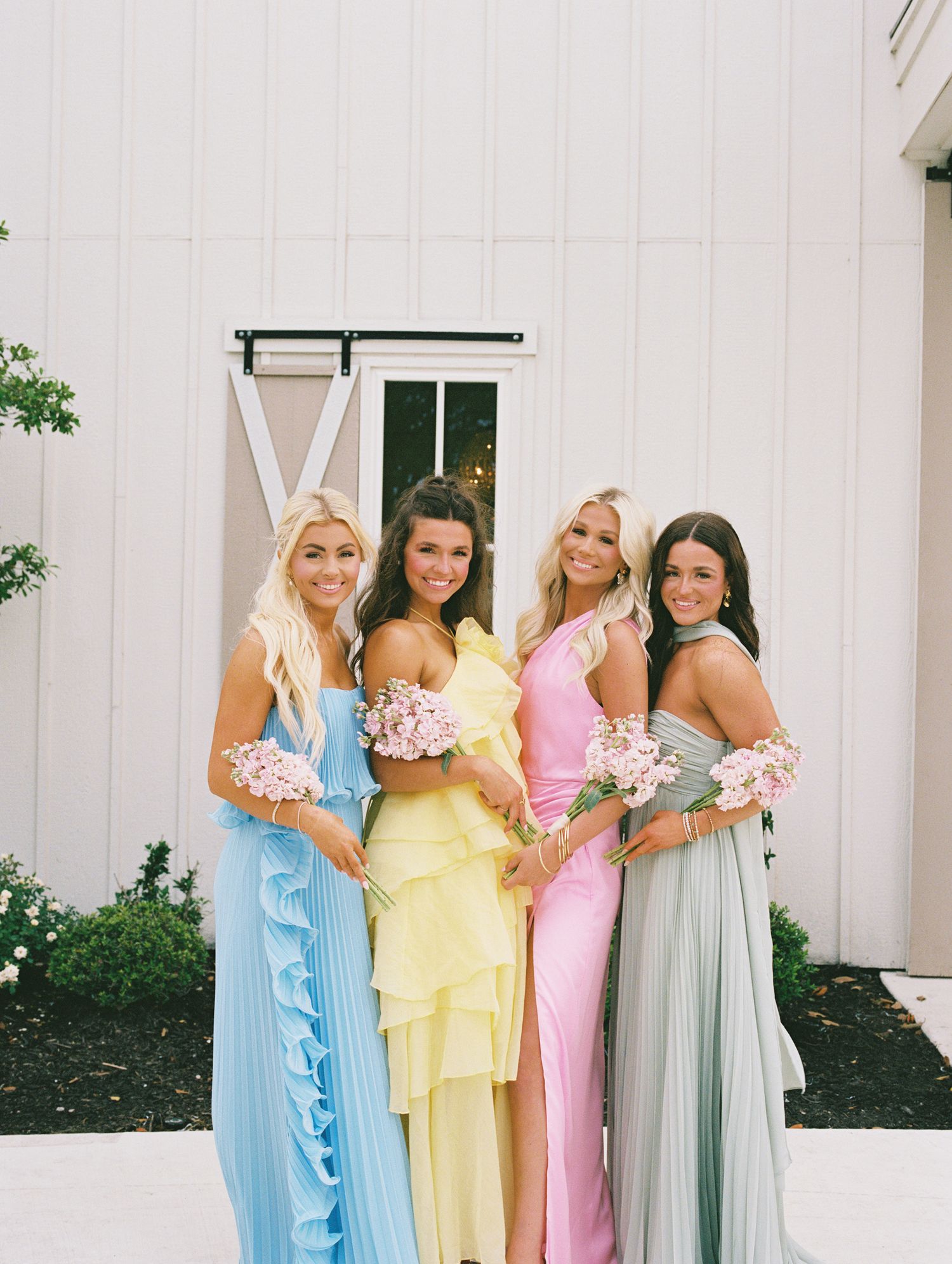 Four bridesmaids pose in pastel gowns holding pink bouquets outside white barn wedding venue.