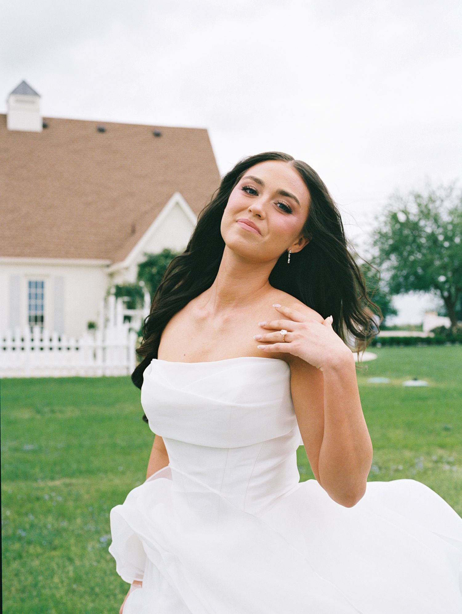 Bride in white strapless gown twirling outdoors near white chapel, dark hair flowing, wearing ring, lush green lawn.