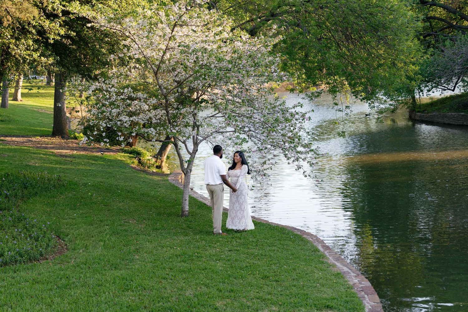 Couple in white attire standing by a serene pond under blooming trees in a lush green park.