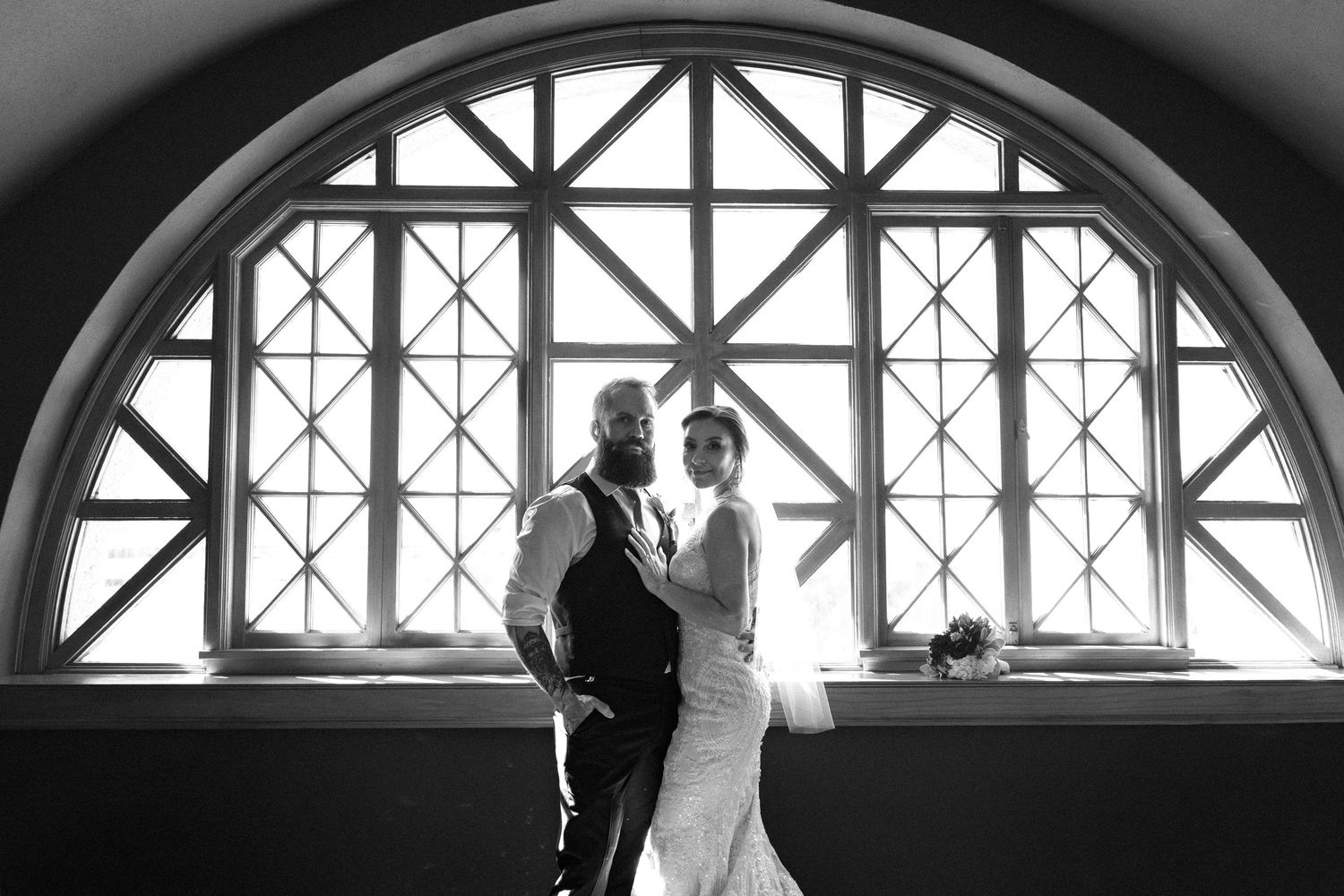 Black and white wedding photo of bride and groom silhouetted against a grand arched window.