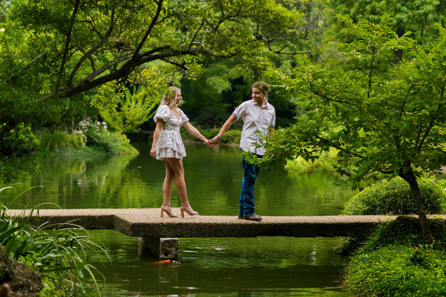 People walking and relaxing on a wooden boardwalk over a tranquil pond surrounded by green summer foliage.