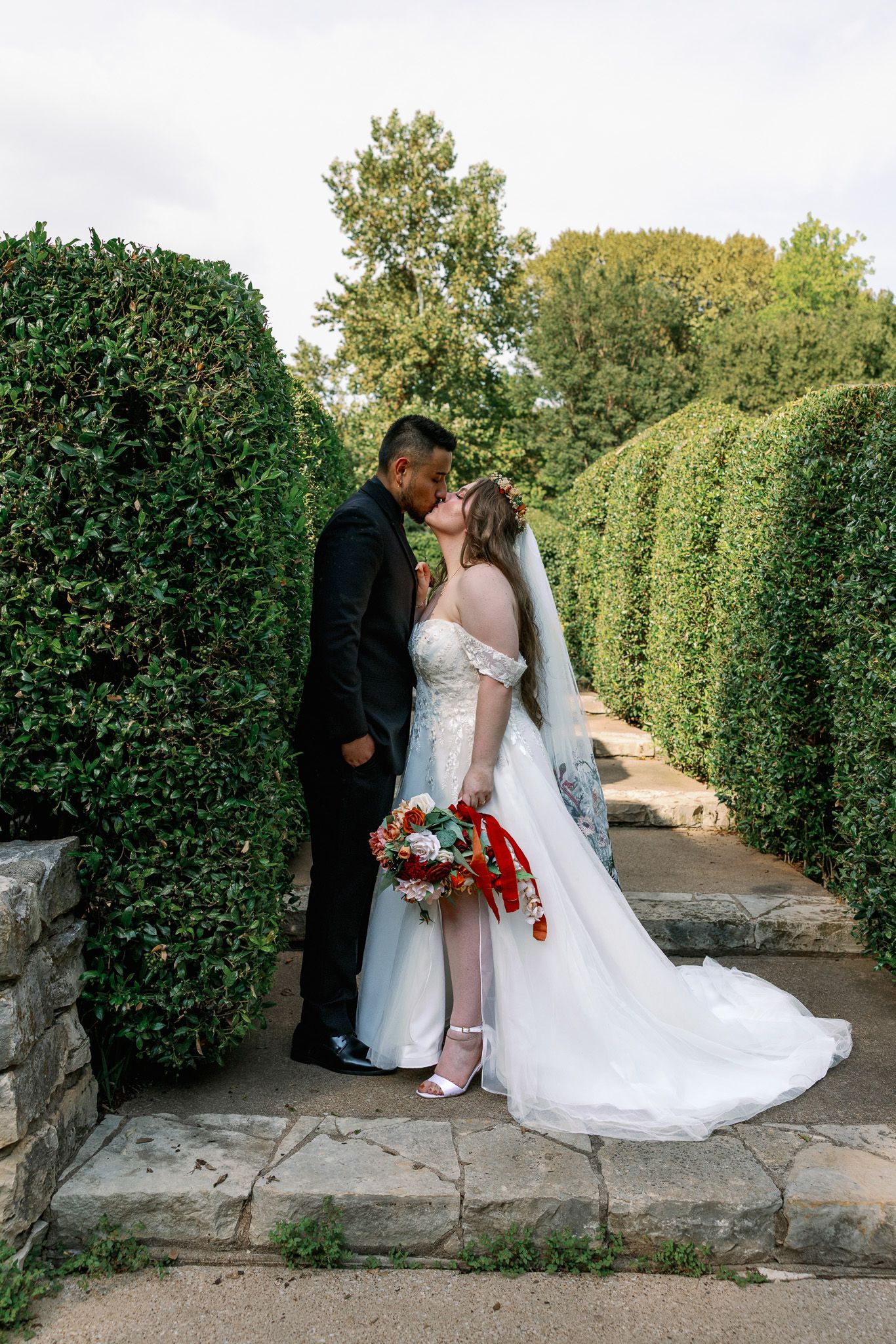 Wedding couple poses along stone wall with manicured hedges in garden setting.
