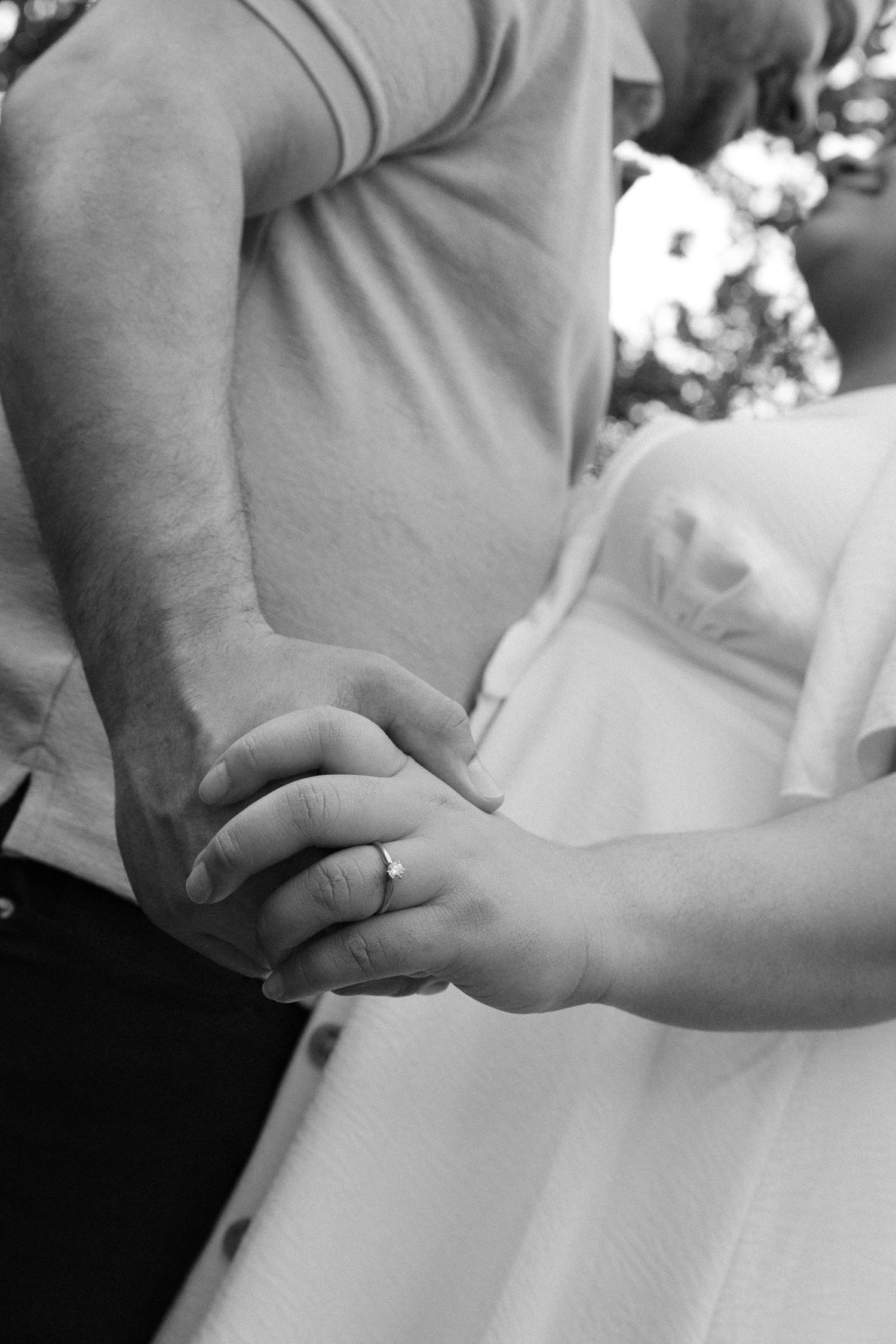 Black and white photo of a couple holding hands, bride wearing wedding ring in white dress with bouquet in background.