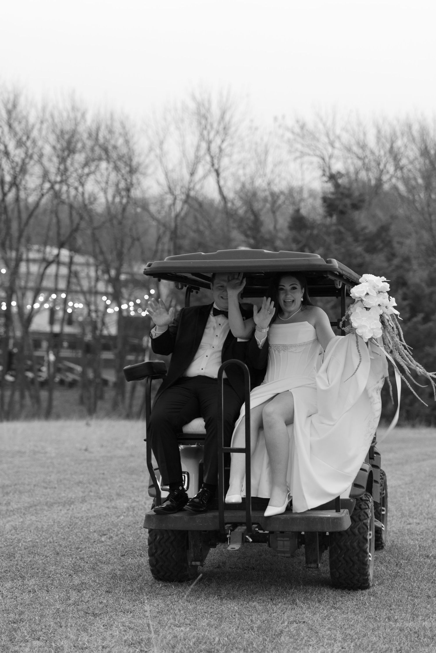 Bride and groom riding a golf cart outdoors on their wedding day, black and white photo with floral bouquet.