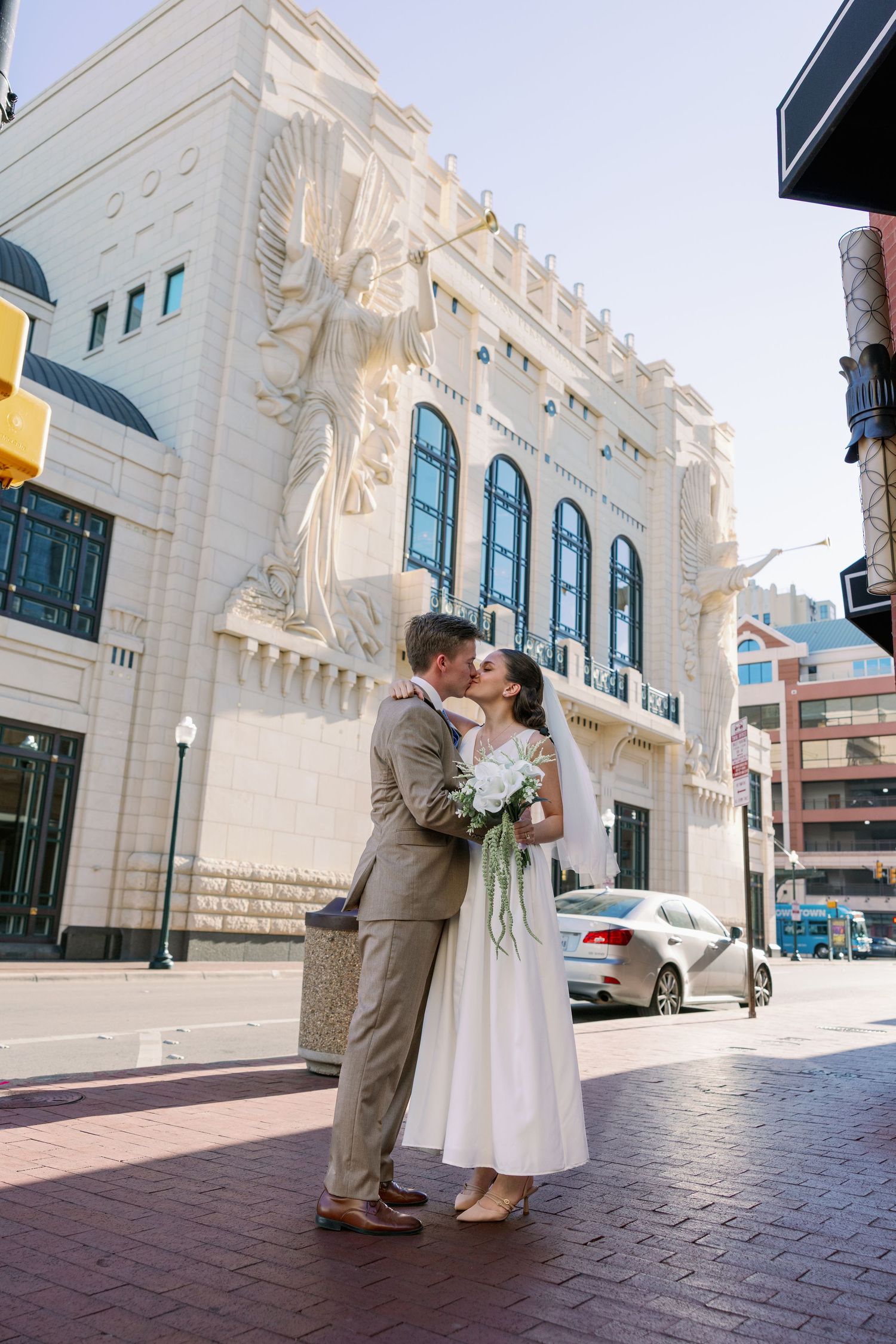 Newlyweds share a kiss on a sunny city street, bride holding white bouquet.