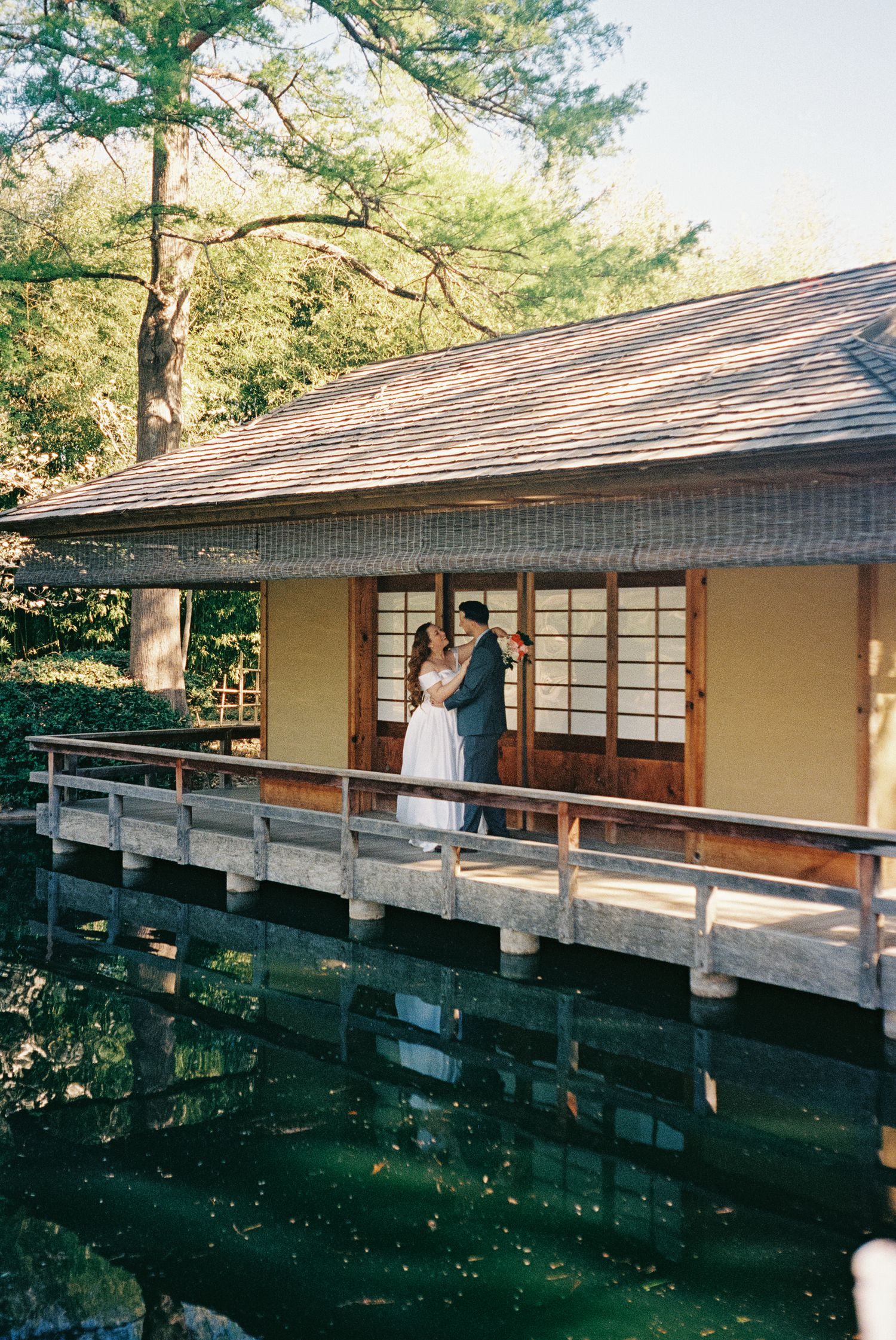 Couple embracing on a serene Japanese tea house deck over a tranquil pond, surrounded by lush greenery.