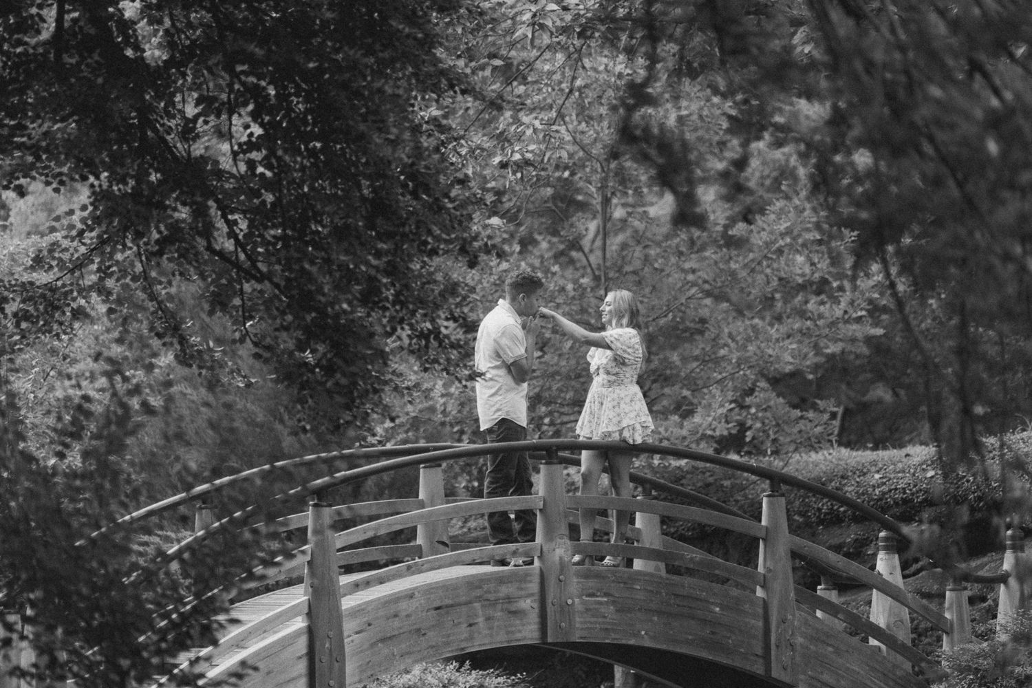 Two figures in white dresses stroll along a wooden bridge path through a lush garden setting.