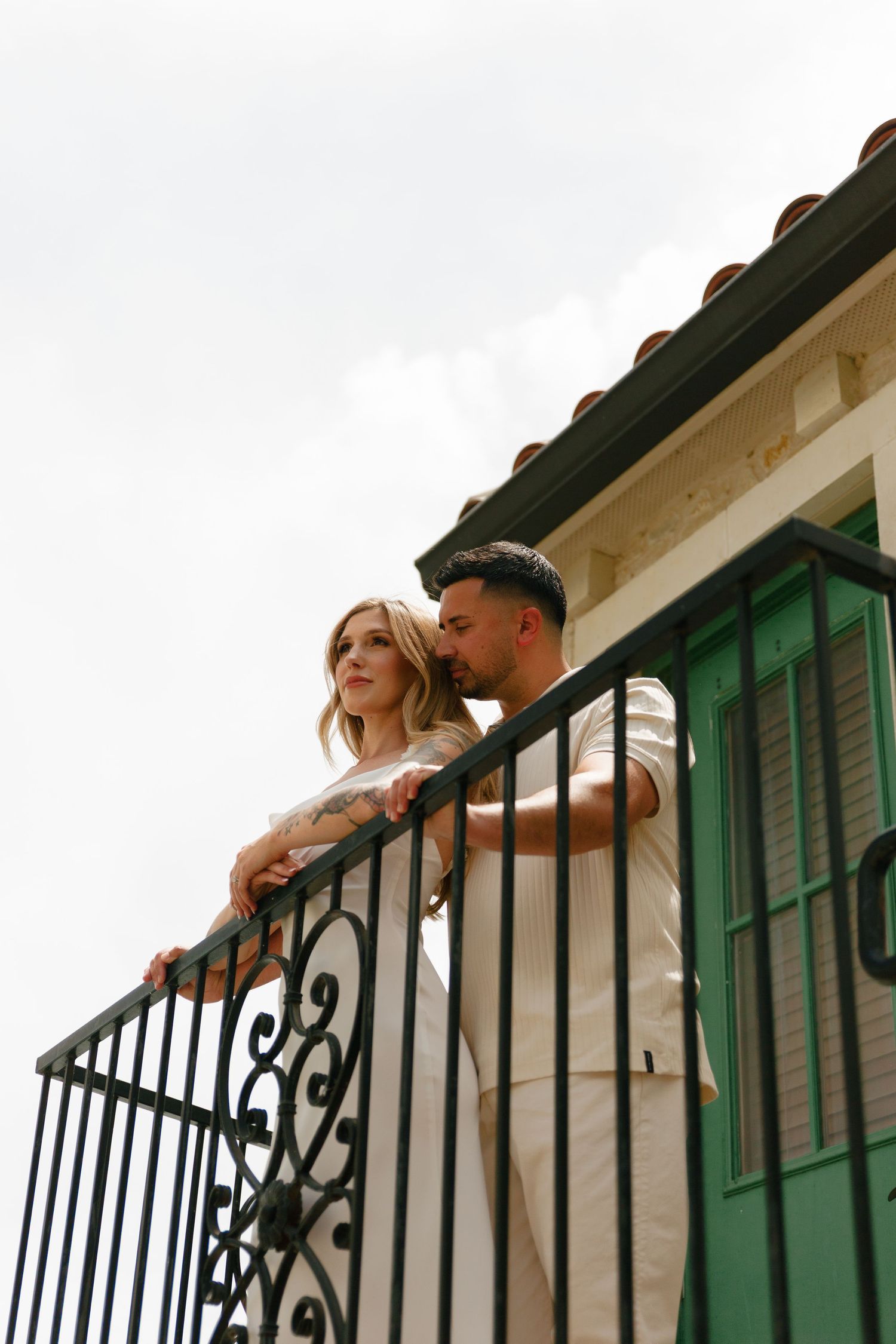 A couple in wedding attire leans against an ornate black railing with green shuttered windows in the background.