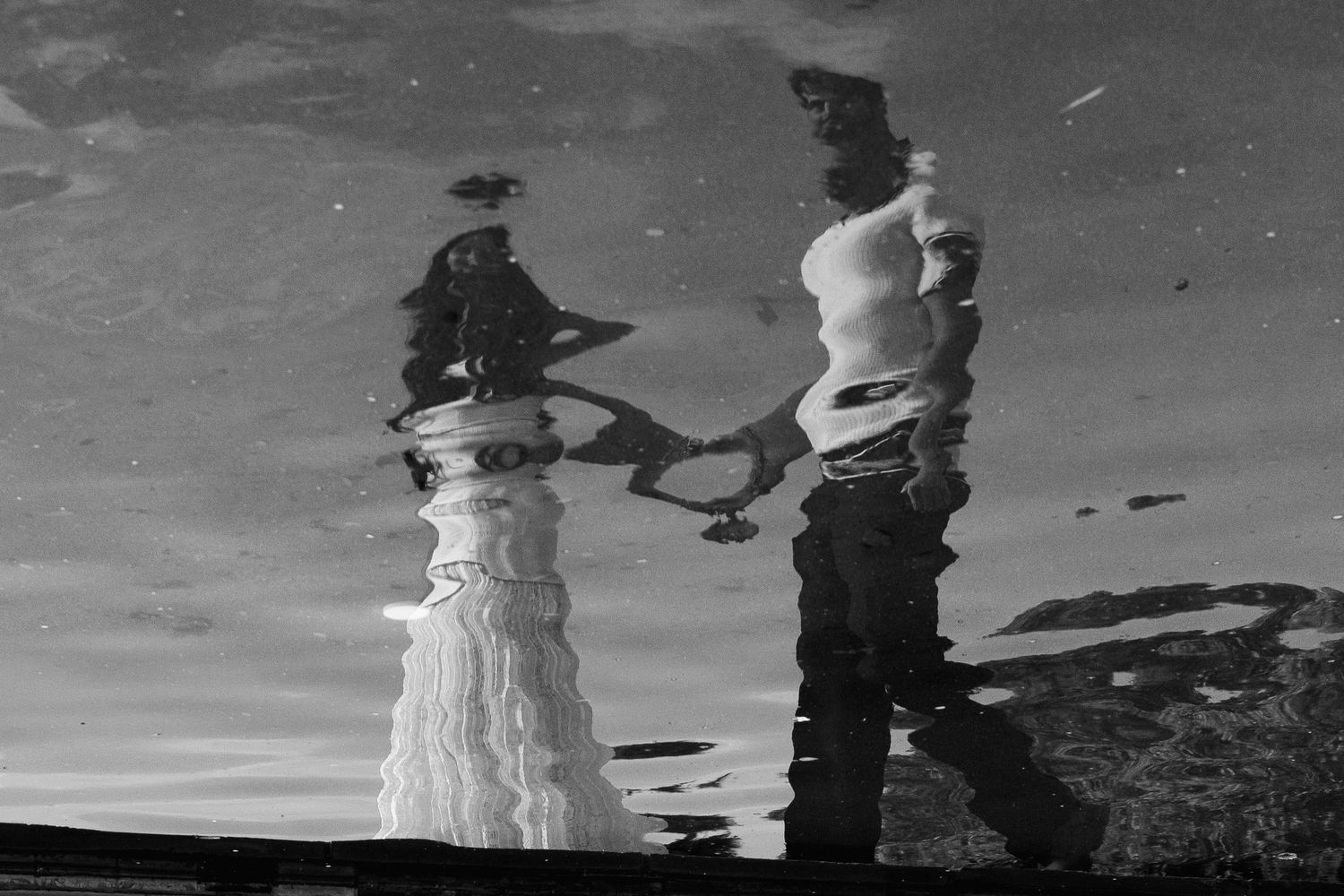 Black and white reflection of a couple holding hands in a puddle, creating an artistic distorted mirror image.