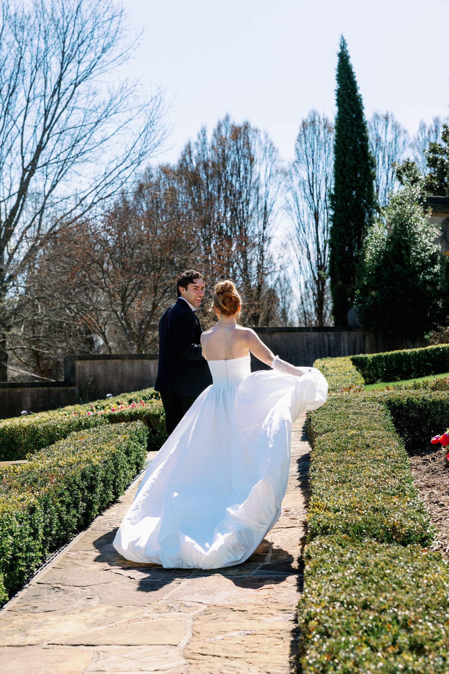 Bride in flowing white gown twirls with groom on garden path lined with manicured hedges and cypress trees.