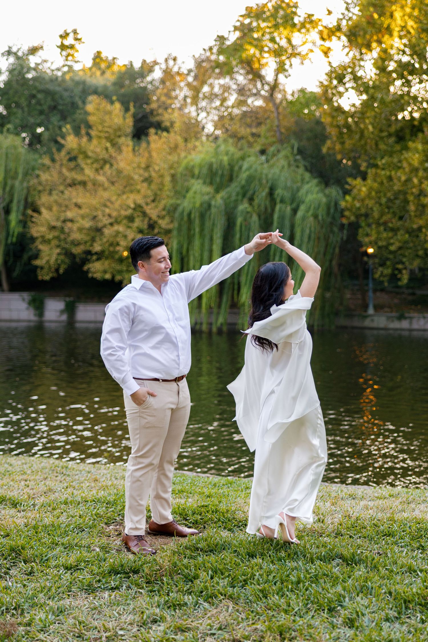 Couple dancing outdoors by a pond, woman in white dress spinning, man in white shirt, lush green trees background.
