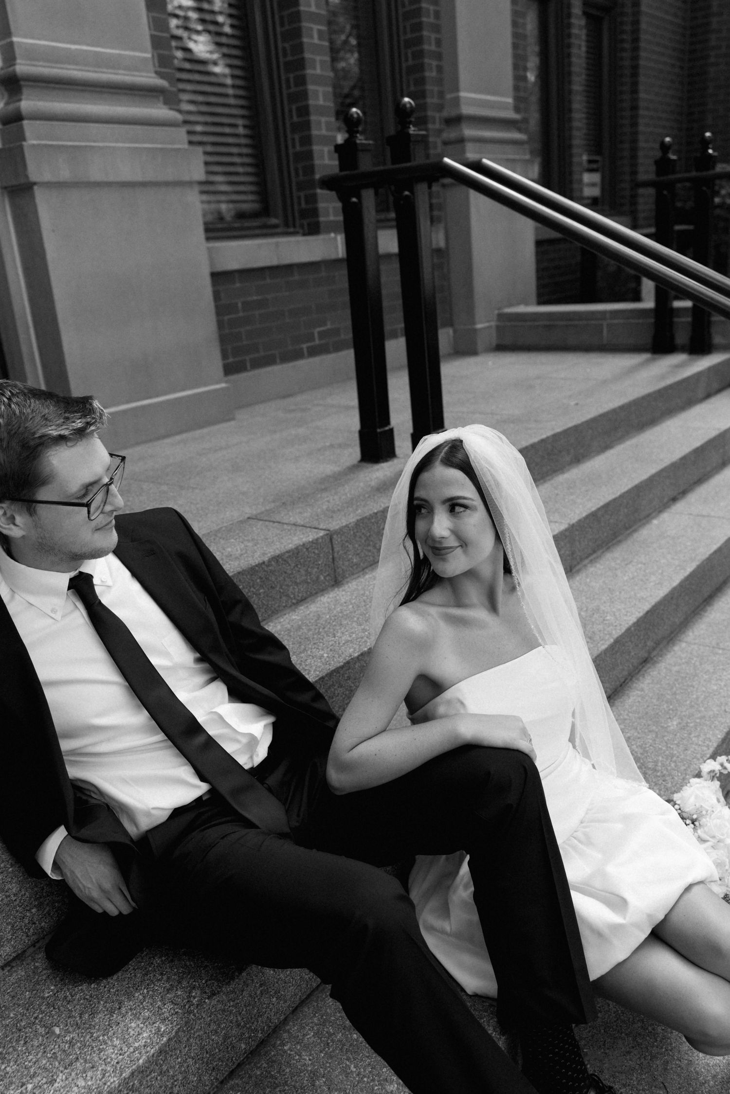 Black and white photo of a bride and groom sitting on church steps, sharing a tender moment in wedding attire.
