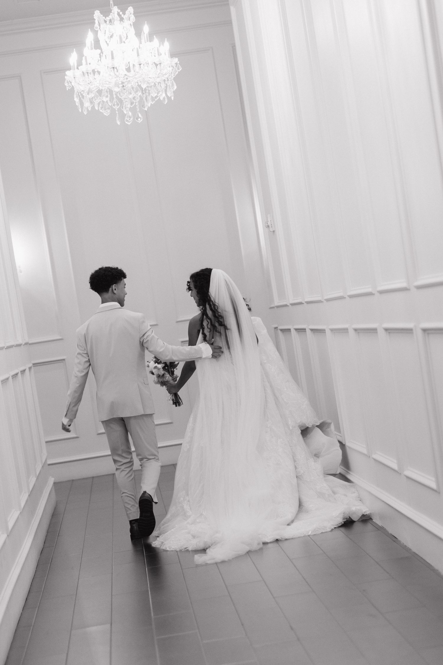 Black and white photo of bride and groom walking hand in hand under crystal chandelier in elegant white hallway.