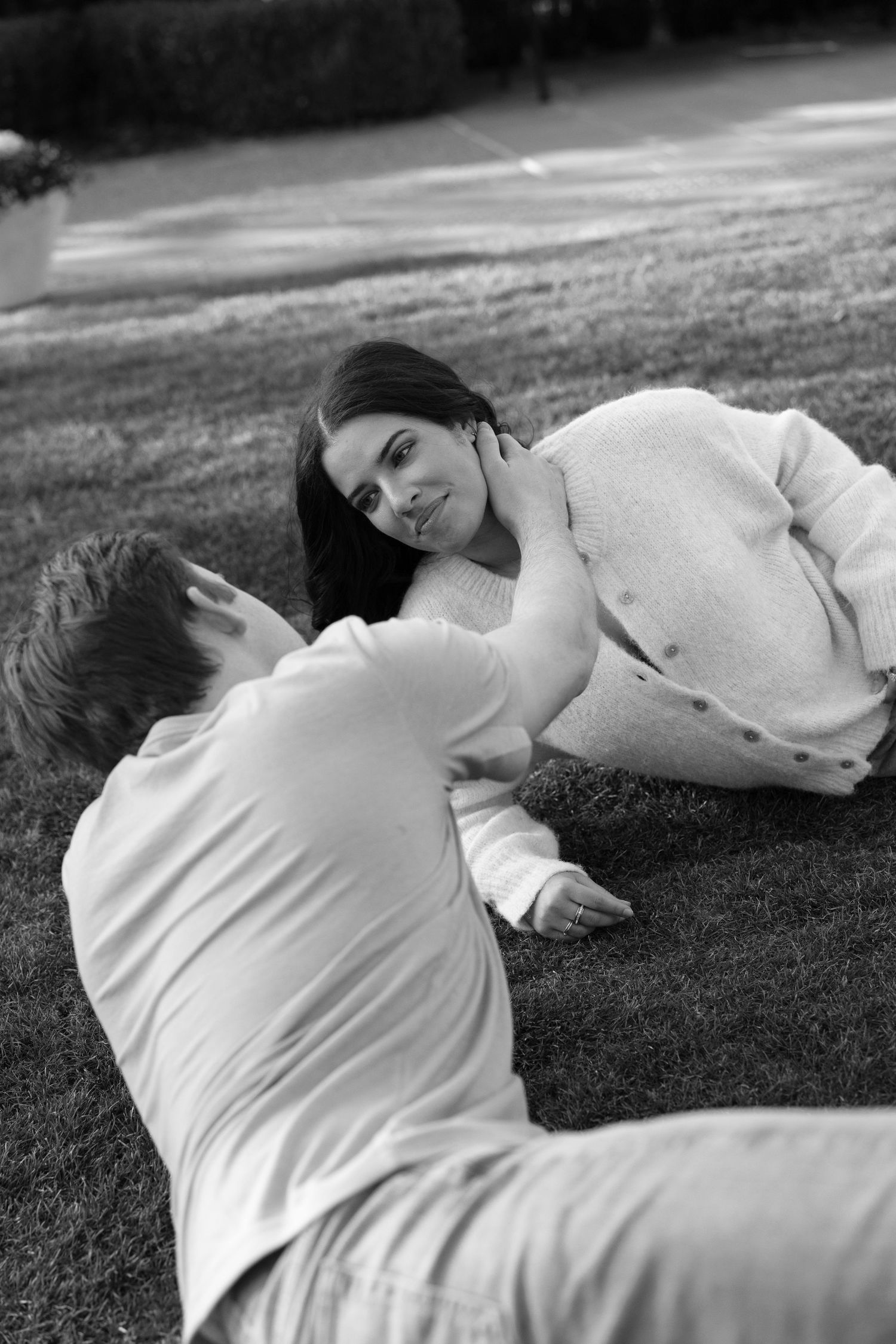 Black and white photo of a couple lying on grass, smiling at each other in a romantic outdoor setting.