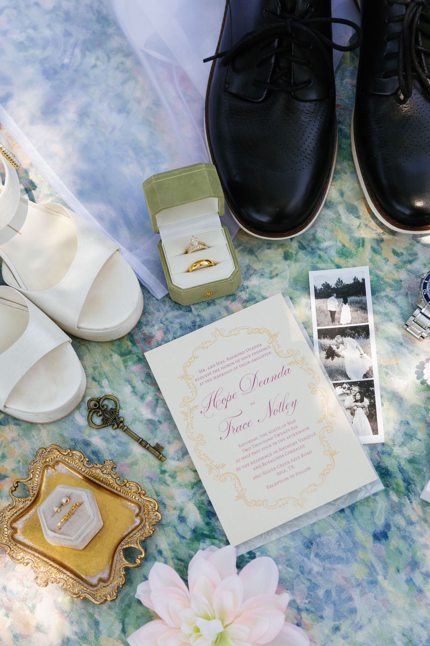 Wedding flat lay featuring rings in a green box, white heels, black dress shoes, invitation, and vintage key on floral fabric.