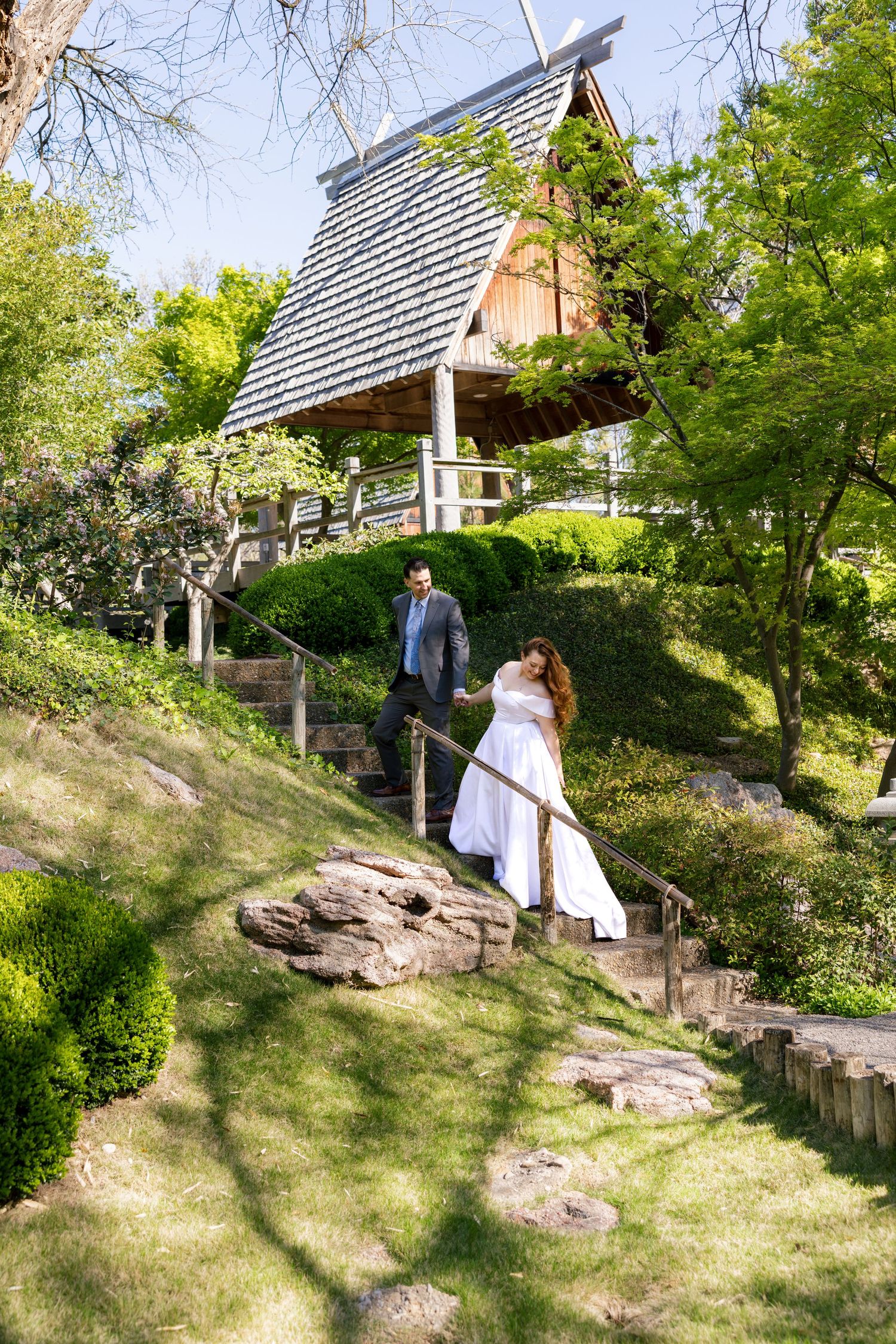 Bride and groom walking up stone steps in a lush Japanese garden with a wooden gazebo in the background.