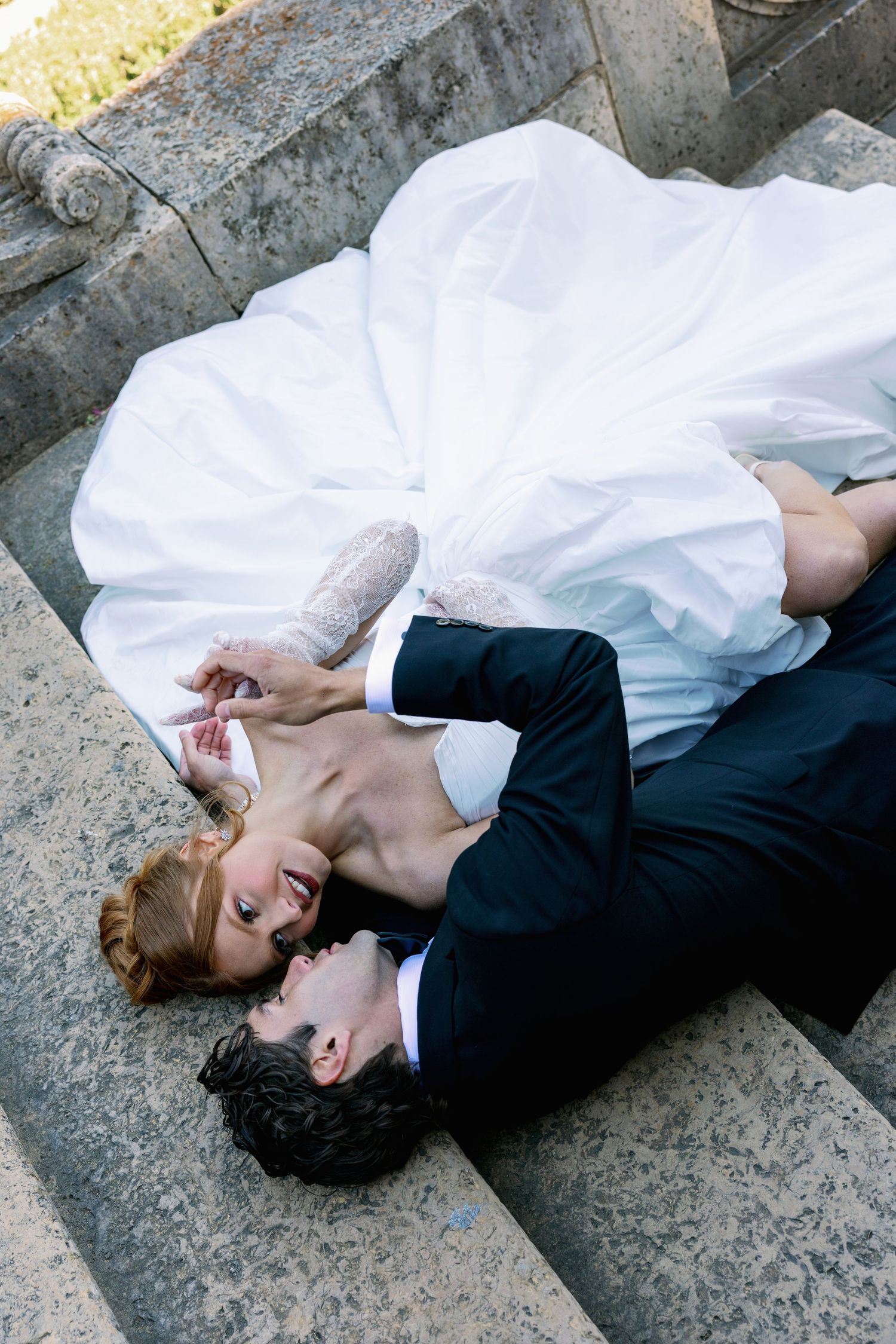 Bride and groom in wedding attire lying on stone steps, sharing an intimate romantic moment.