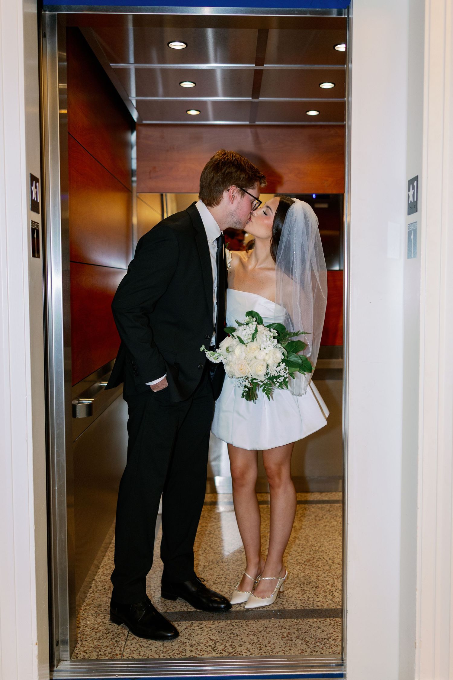 Bride and groom share a kiss in an elevator, bride wearing short white dress with veil holding white bouquet.