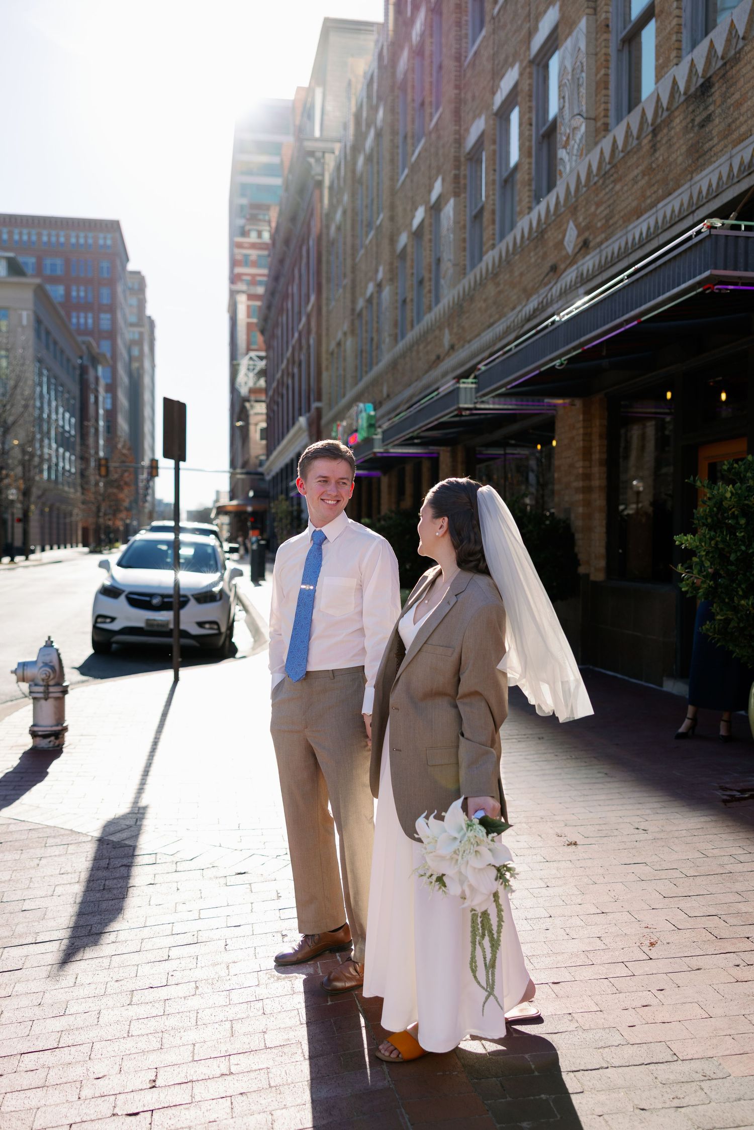 Bride and groom stand on a sunny urban sidewalk, she holds white flowers and wears a veil, he wears a tan suit with blue tie.