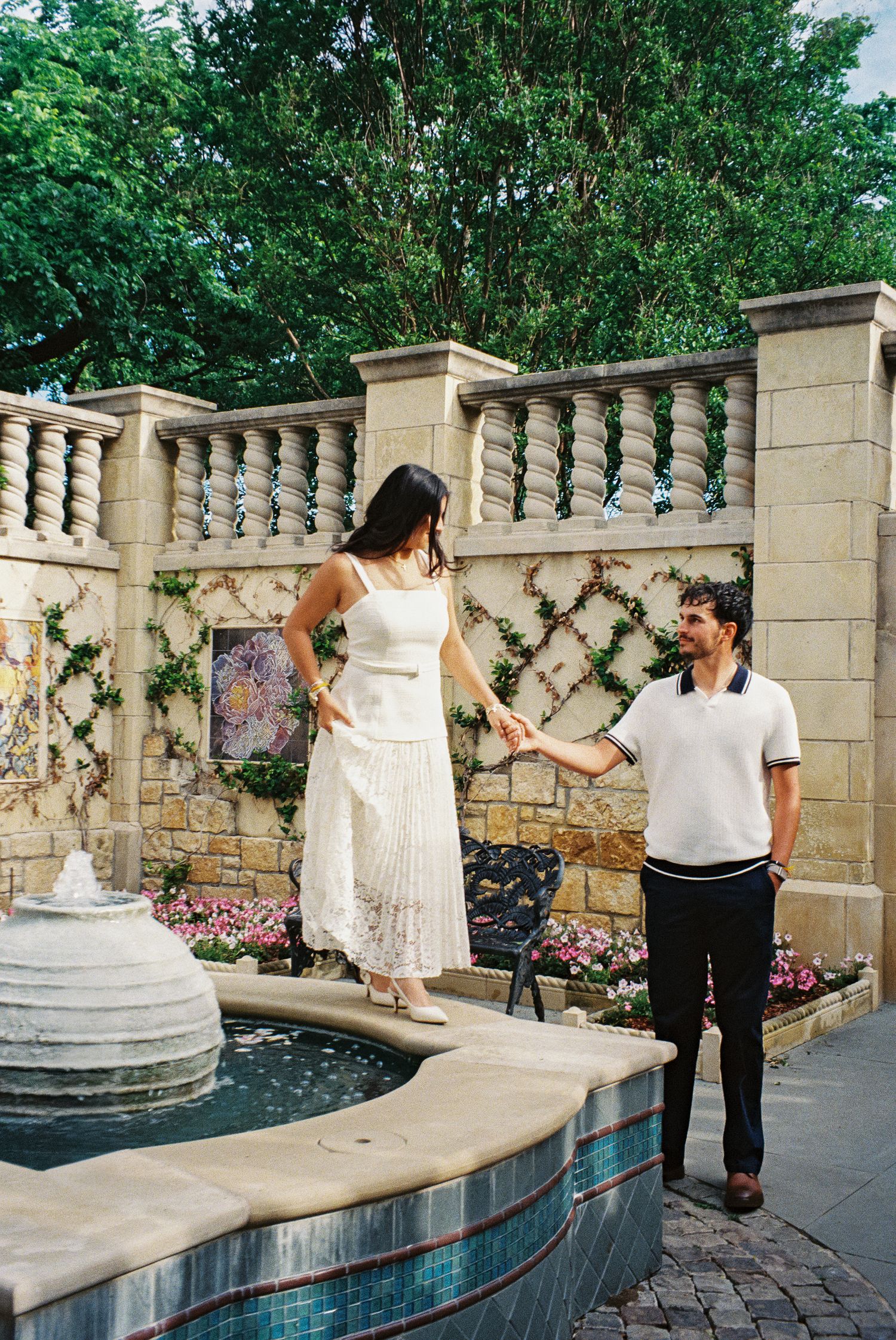 Couple holding hands by an ornate stone fountain in a garden, woman in white dress balancing on edge.