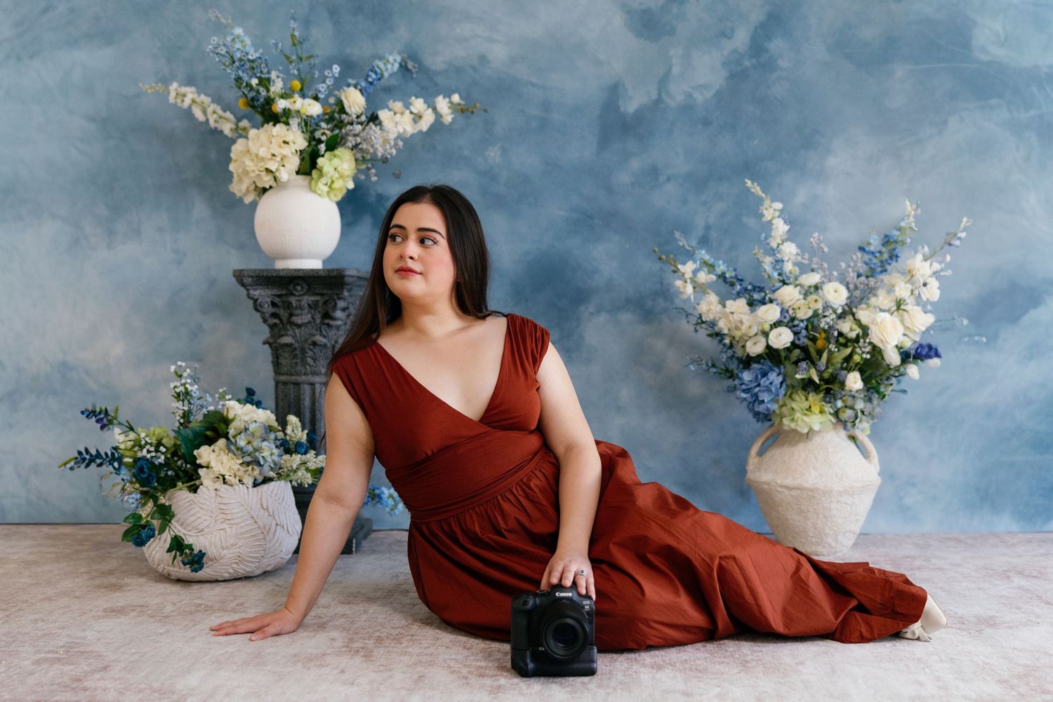 A figure in a burgundy dress poses elegantly against a moody blue backdrop with white floral arrangements.