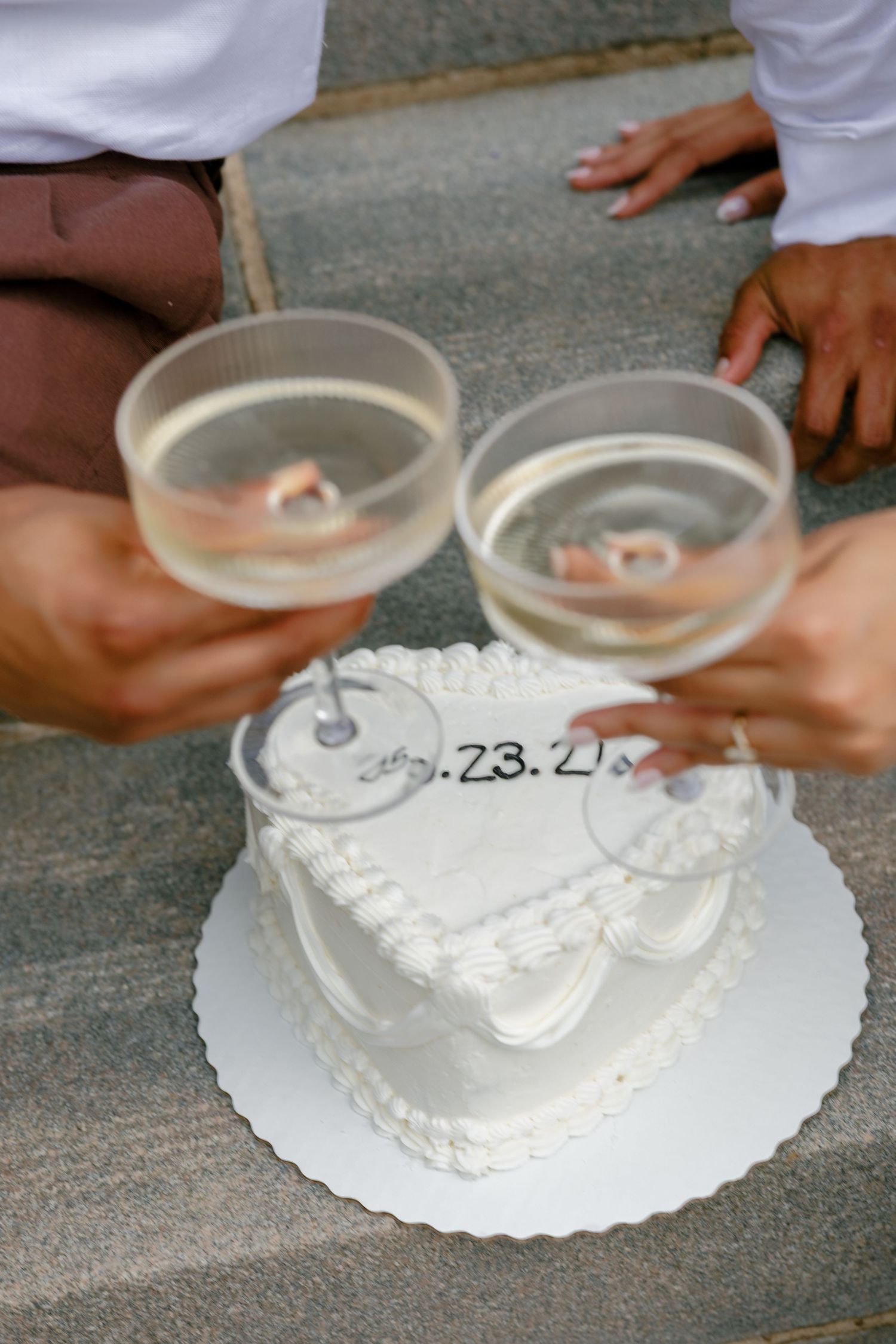 Two hands hold champagne glasses for a toast over a white frosted heart-shaped wedding cake with date writing.