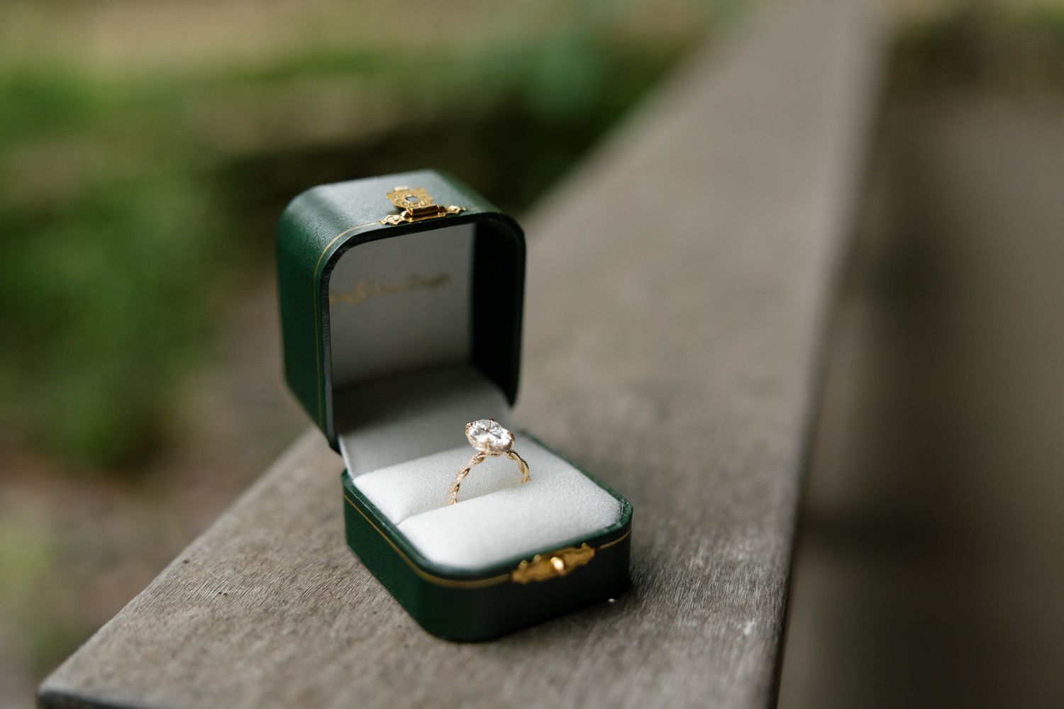 A sparkling diamond engagement ring sits in an open dark green velvet ring box on a stone surface.