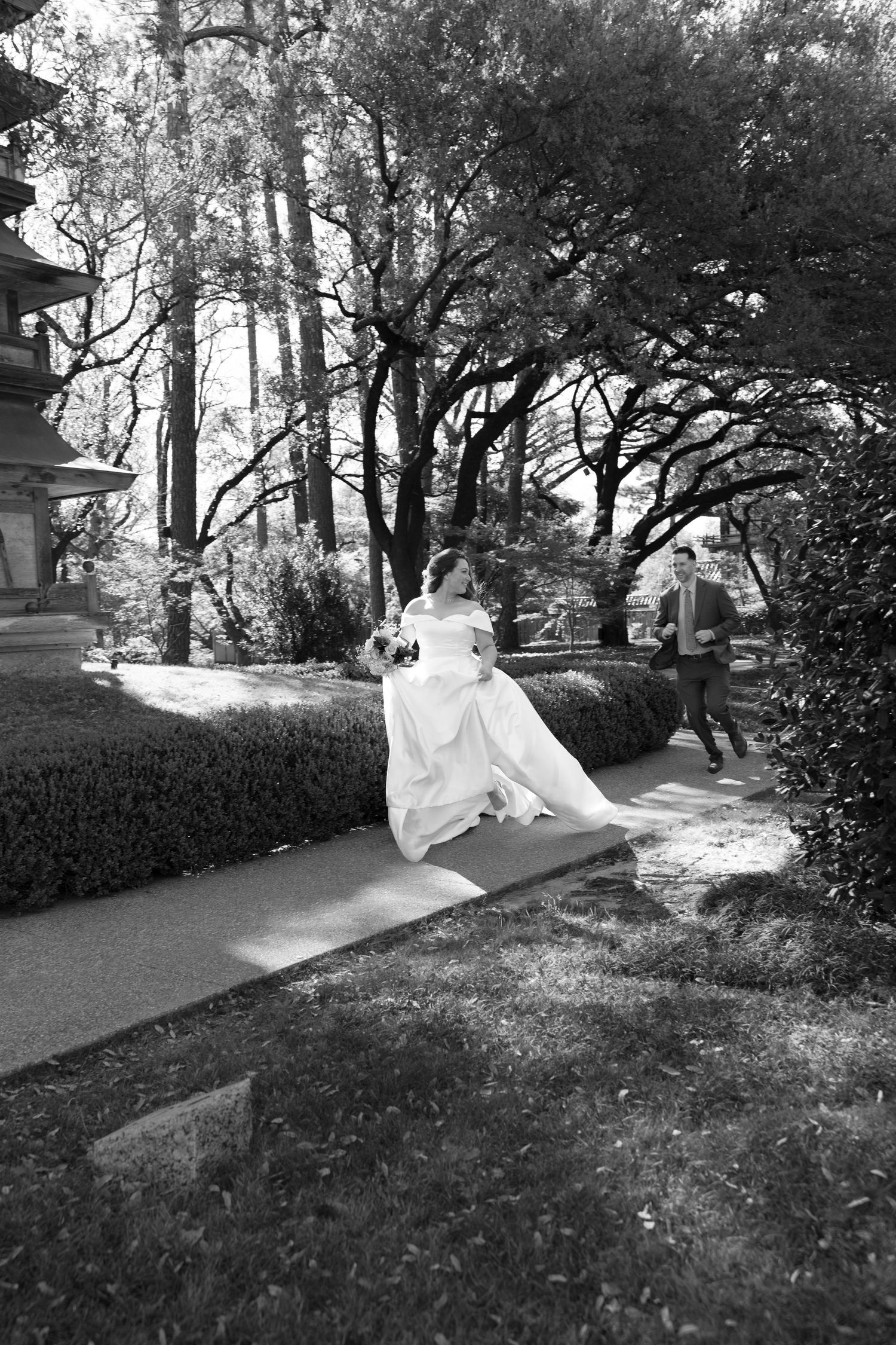 Black and white photo of a bride in a white gown sitting outdoors with a groom approaching on a garden path.