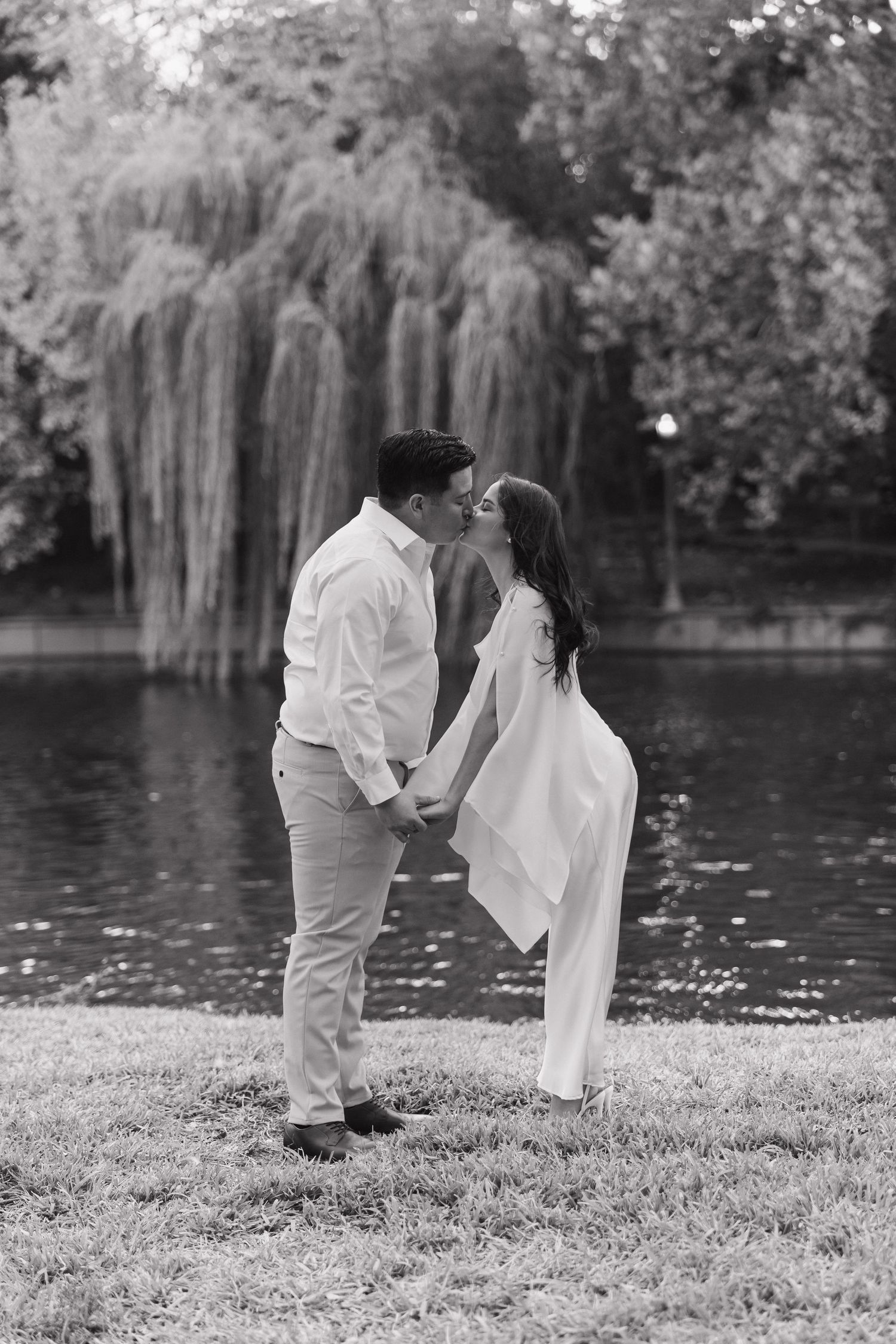 Black and white photo of a romantic couple holding hands and kissing by a tranquil pond with weeping willow trees.